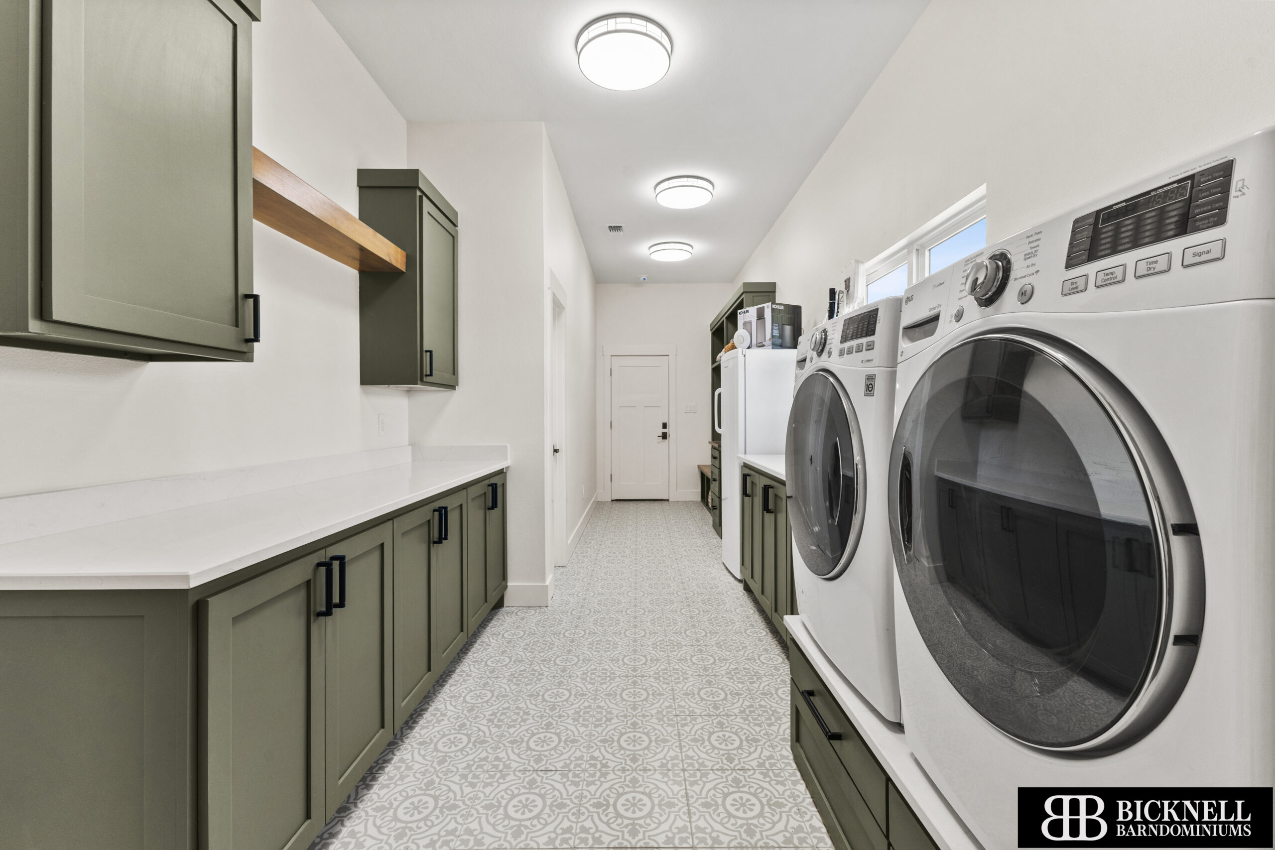 Spacious laundry room with green cabinets, white countertops, front-loading washer and dryer, patterned tile floor, and overhead lighting.