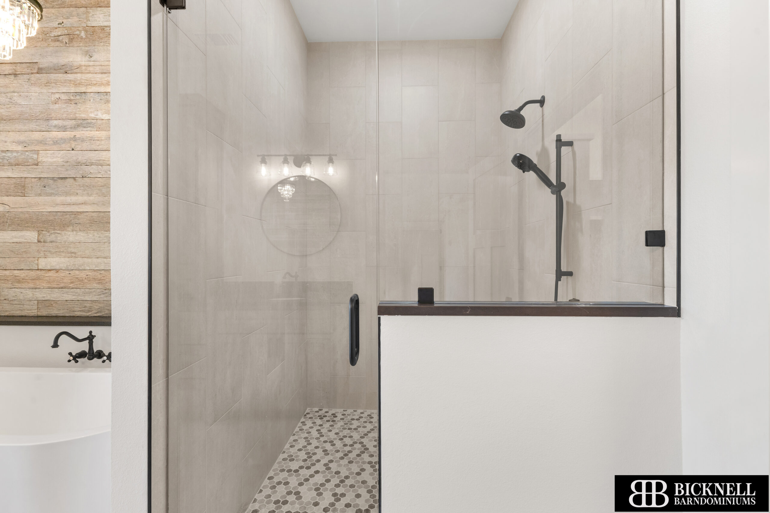 Modern walk-in shower with glass door, beige tile walls, black fixtures, and pebble tile floor. Freestanding bathtub visible on the left. Bicknell logo in bottom right corner.