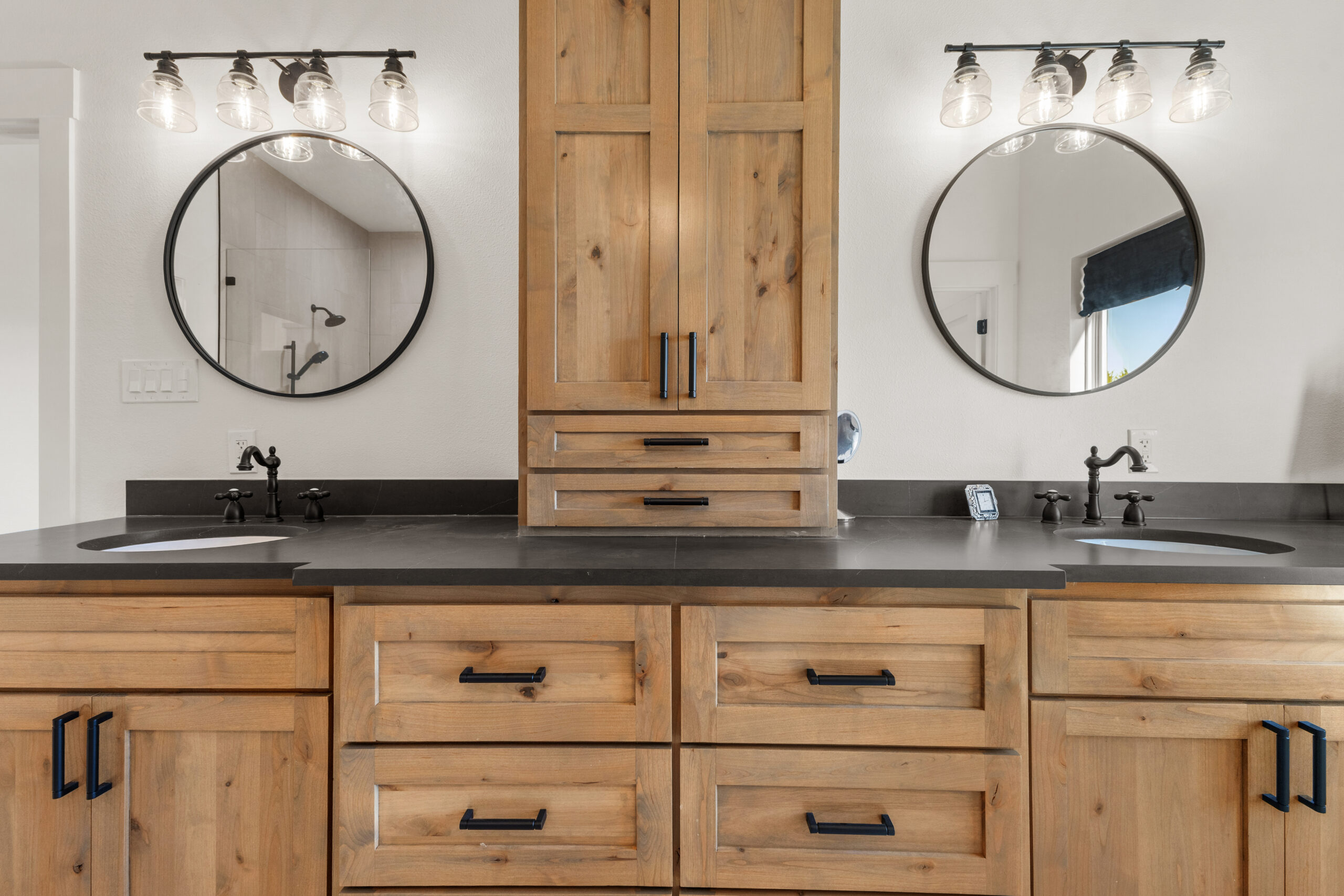 Symmetrical bathroom vanity with wooden cabinets, black countertop, round mirrors, and black faucets.