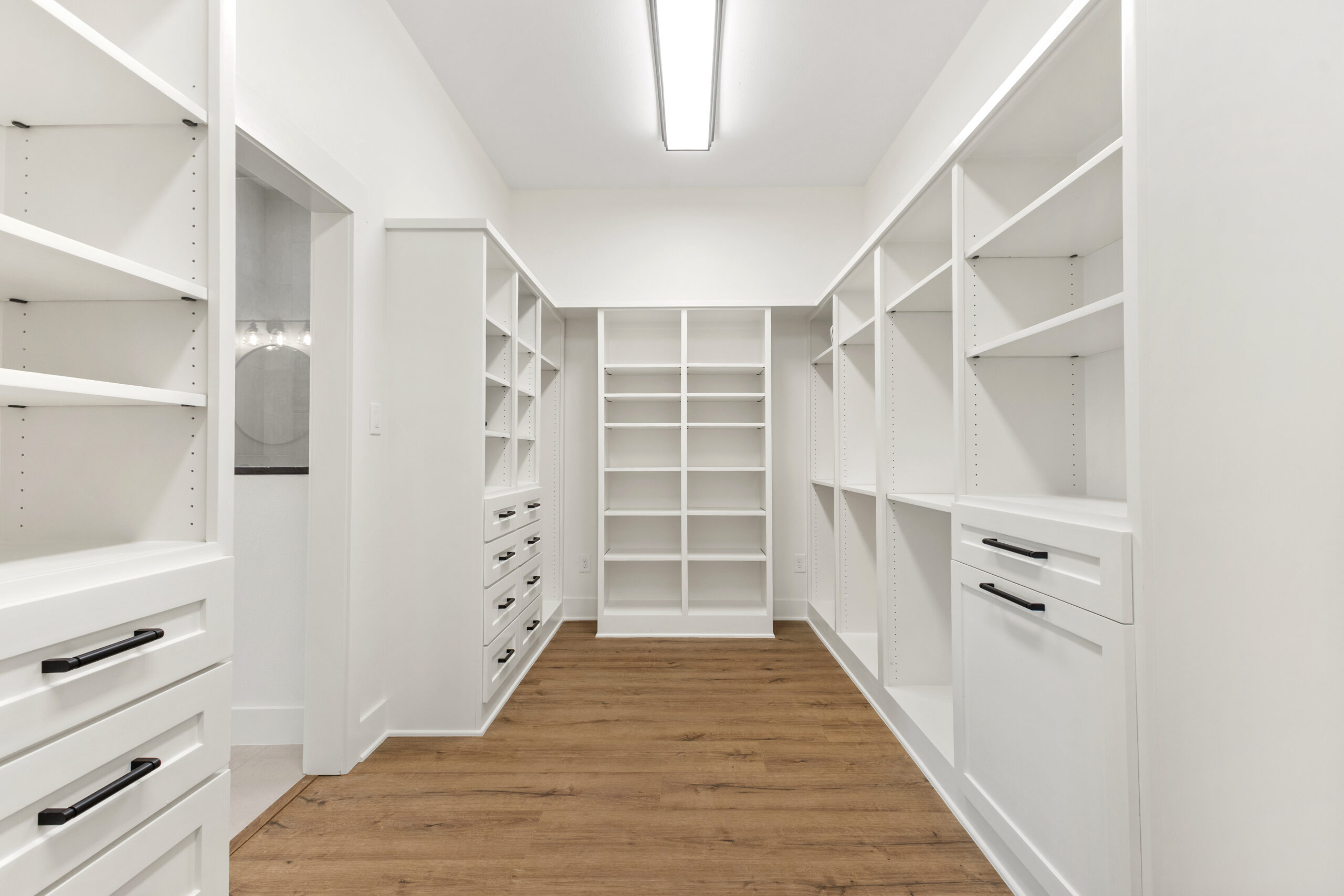 Spacious walk-in closet with built-in white shelves, drawers, and cabinets, featuring a wooden floor and overhead fluorescent lighting.