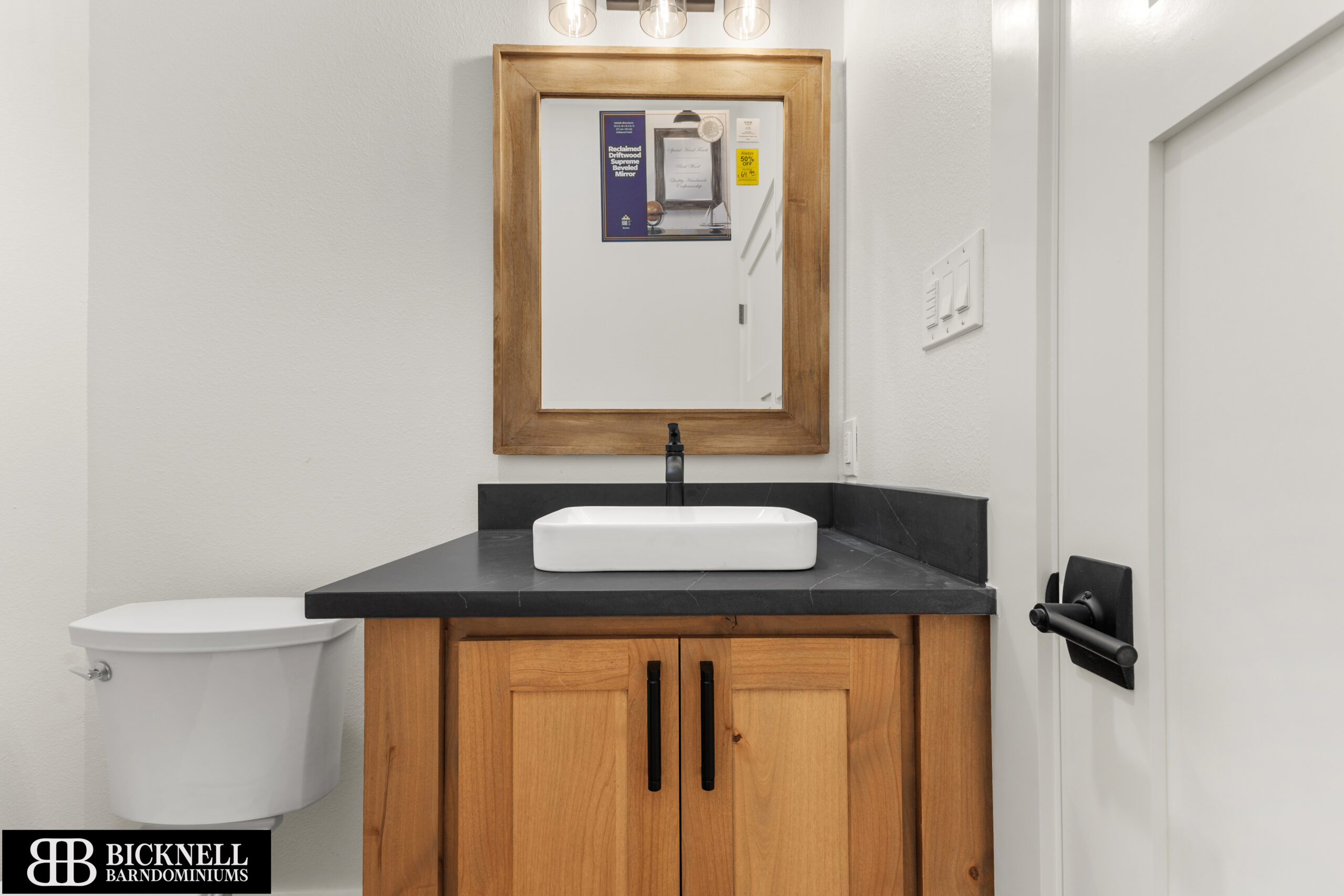 A modern bathroom with a wooden vanity, rectangular mirror, white vessel sink, black faucet, and toilet. A door with a black handle is partially visible on the right.
