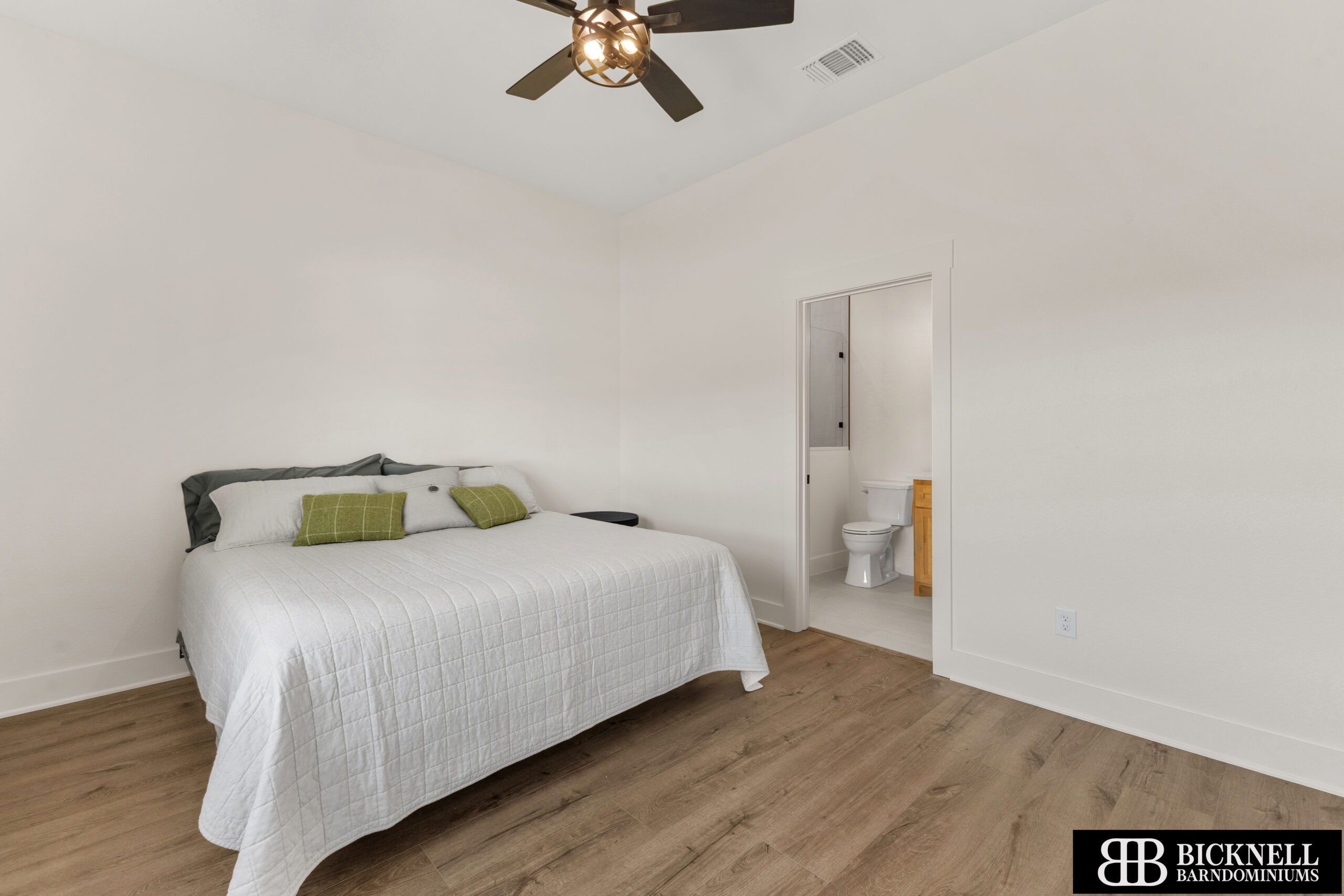 Bedroom with a white quilted bed, green pillows, and wood flooring; open doorway to a small bathroom visible on the right.
