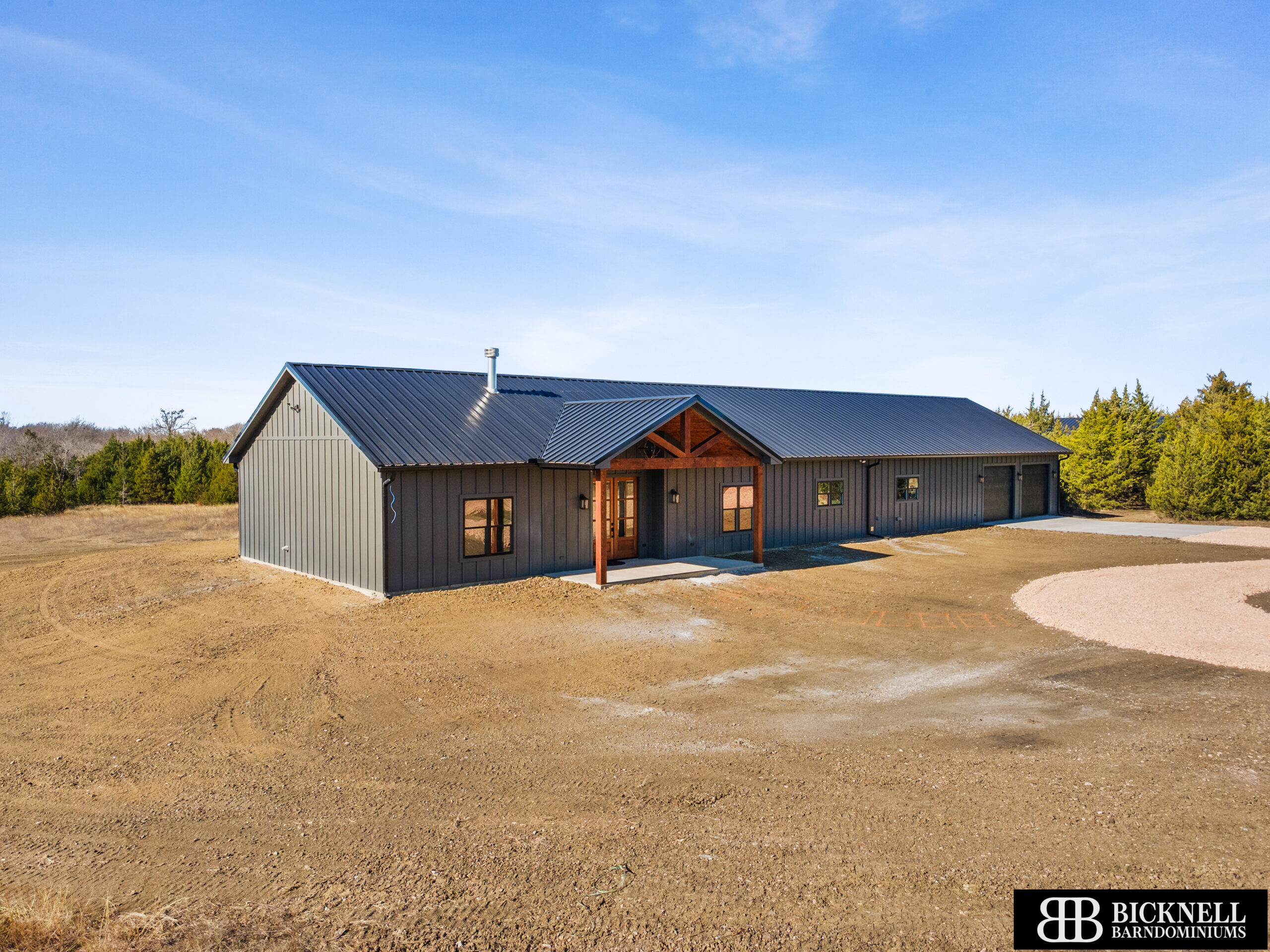 A single-story, metal-clad barndominium with a covered entryway sits on cleared land surrounded by trees under a blue sky.