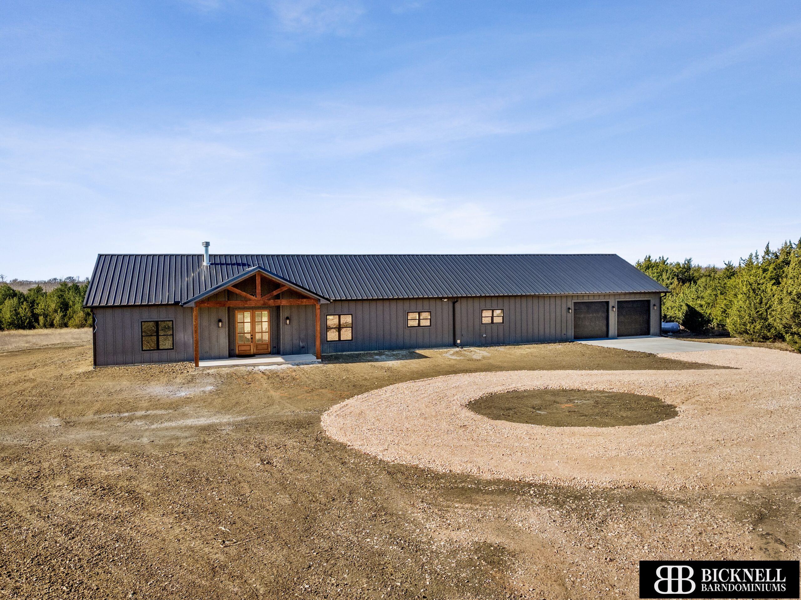 A single-story, metal-sided barndominium with a covered porch, attached garage, and circular gravel driveway, set in a rural landscape under a clear sky.