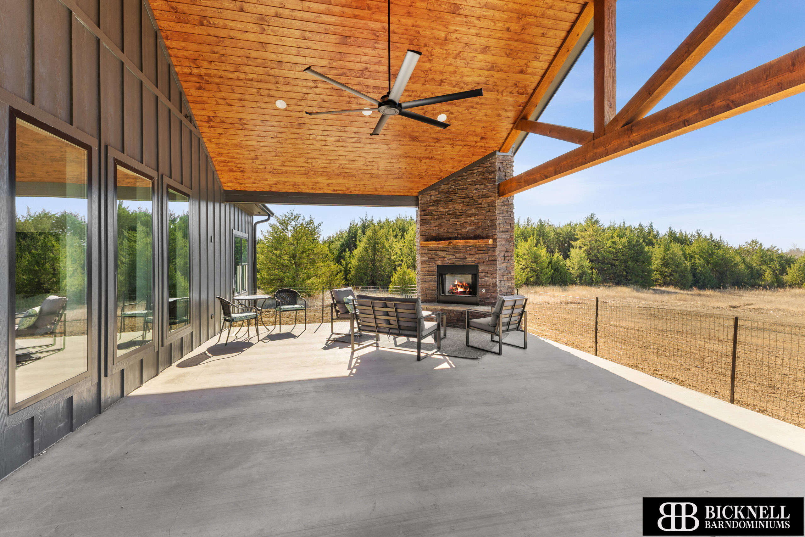 A covered patio with a wood ceiling, ceiling fan, stone fireplace, outdoor seating, and a view of trees; Bicknell Barndominiums logo in the corner.