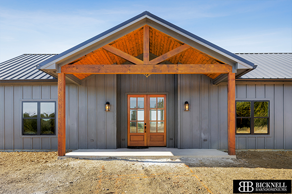 A modern farmhouse with vertical gray siding, a metal roof, and a wooden front entry featuring exposed beams, double doors, and glass panels. Bicknell Barndominiums logo is visible.