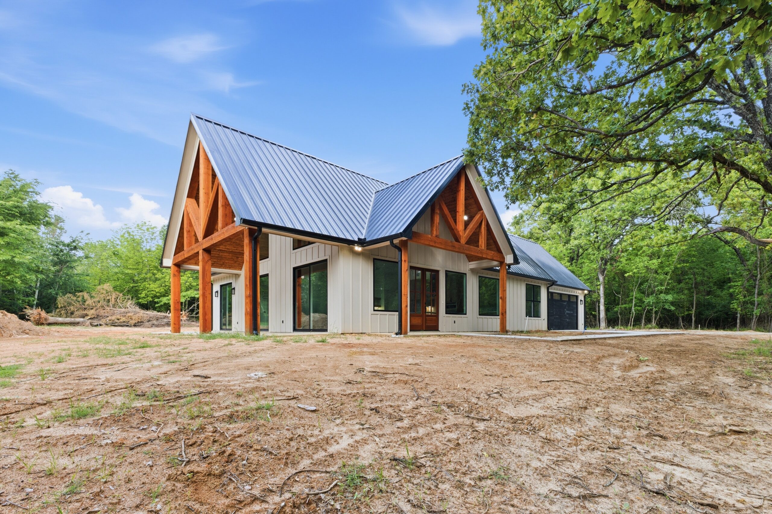Modern house with a steep metal roof, wood and white siding accents, large windows, and a three-car garage, surrounded by trees and open land under a blue sky.