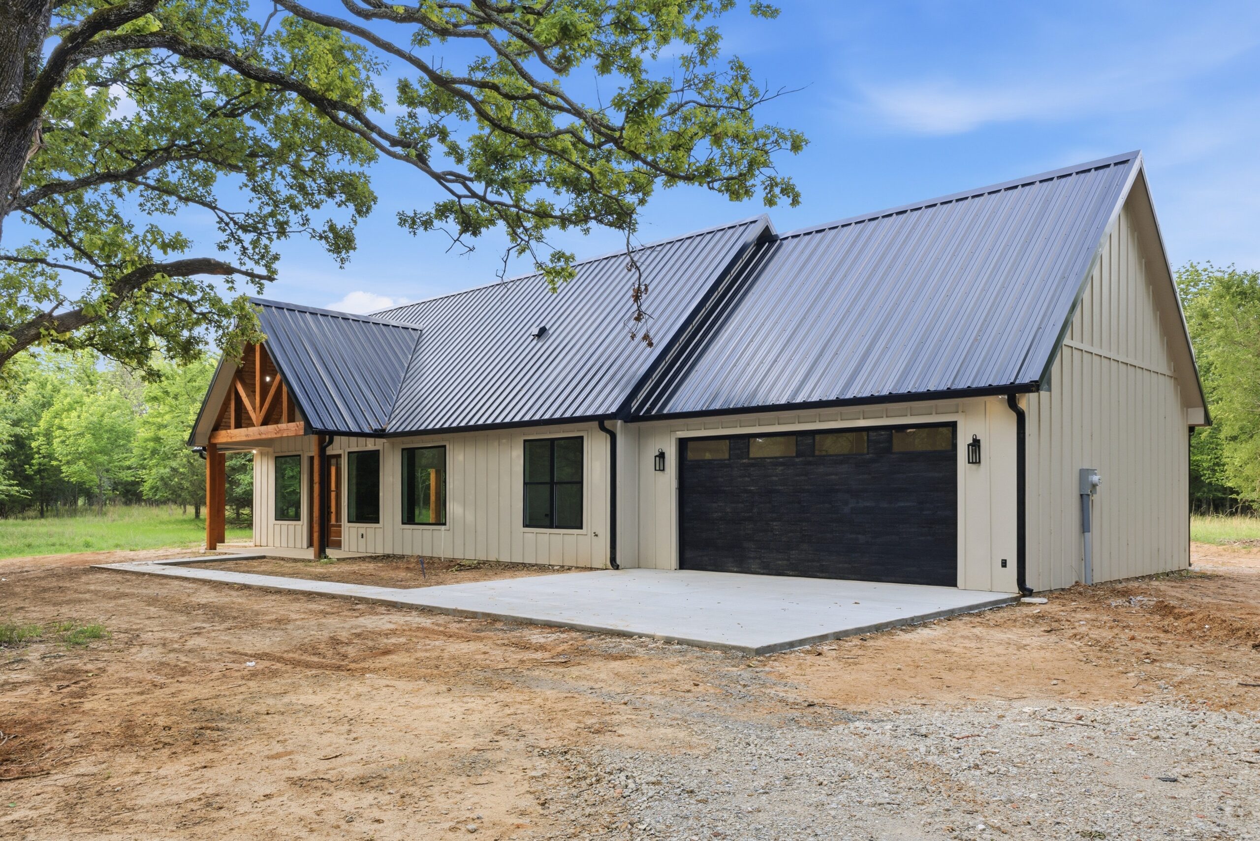 Single-story modern farmhouse with a black metal roof, beige siding, a two-car garage, and a concrete driveway, surrounded by trees and open land.
