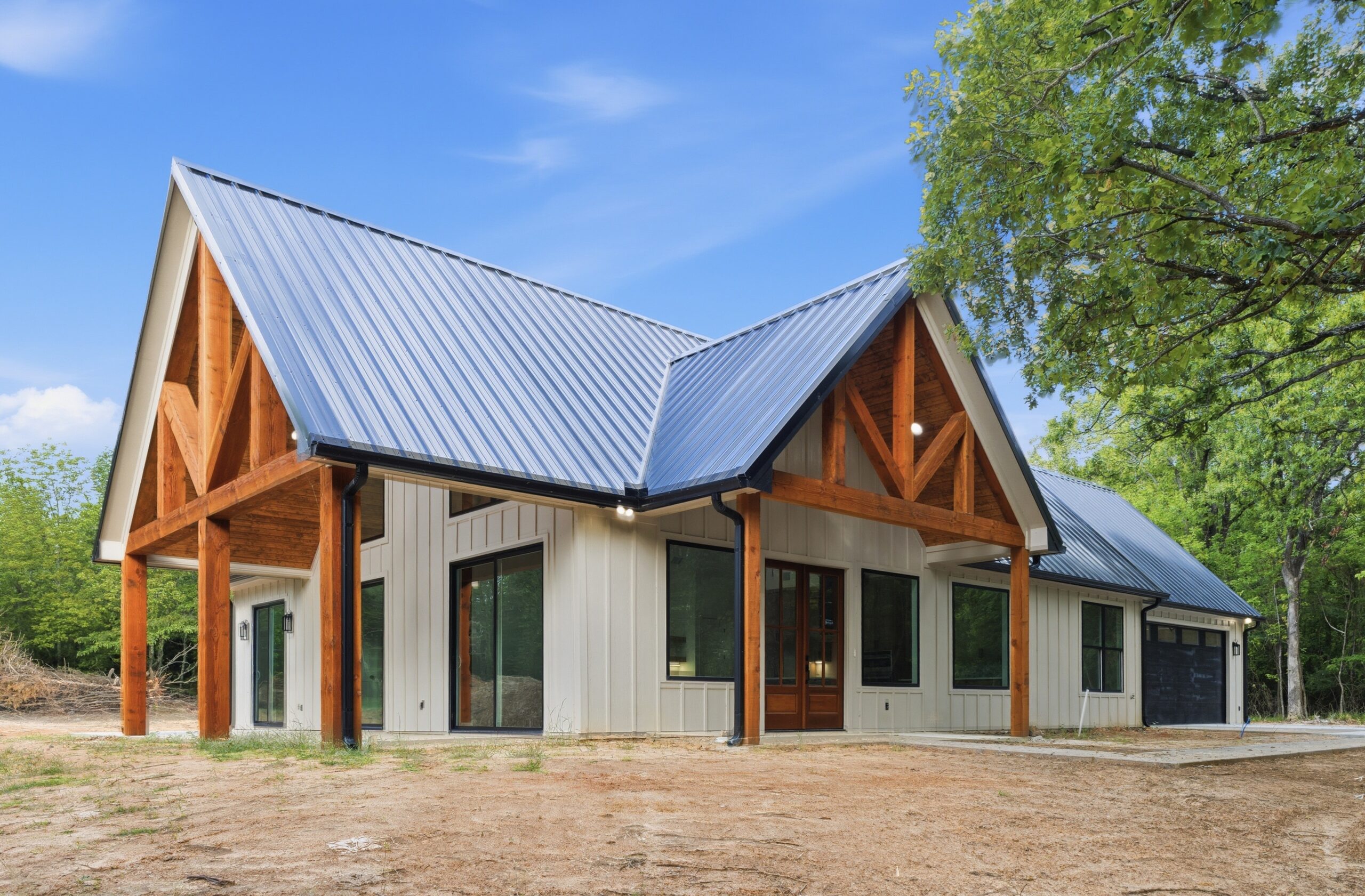 Modern home with a silver metal pitched roof, exposed wooden beams, white siding, and large glass doors/windows under a blue sky.