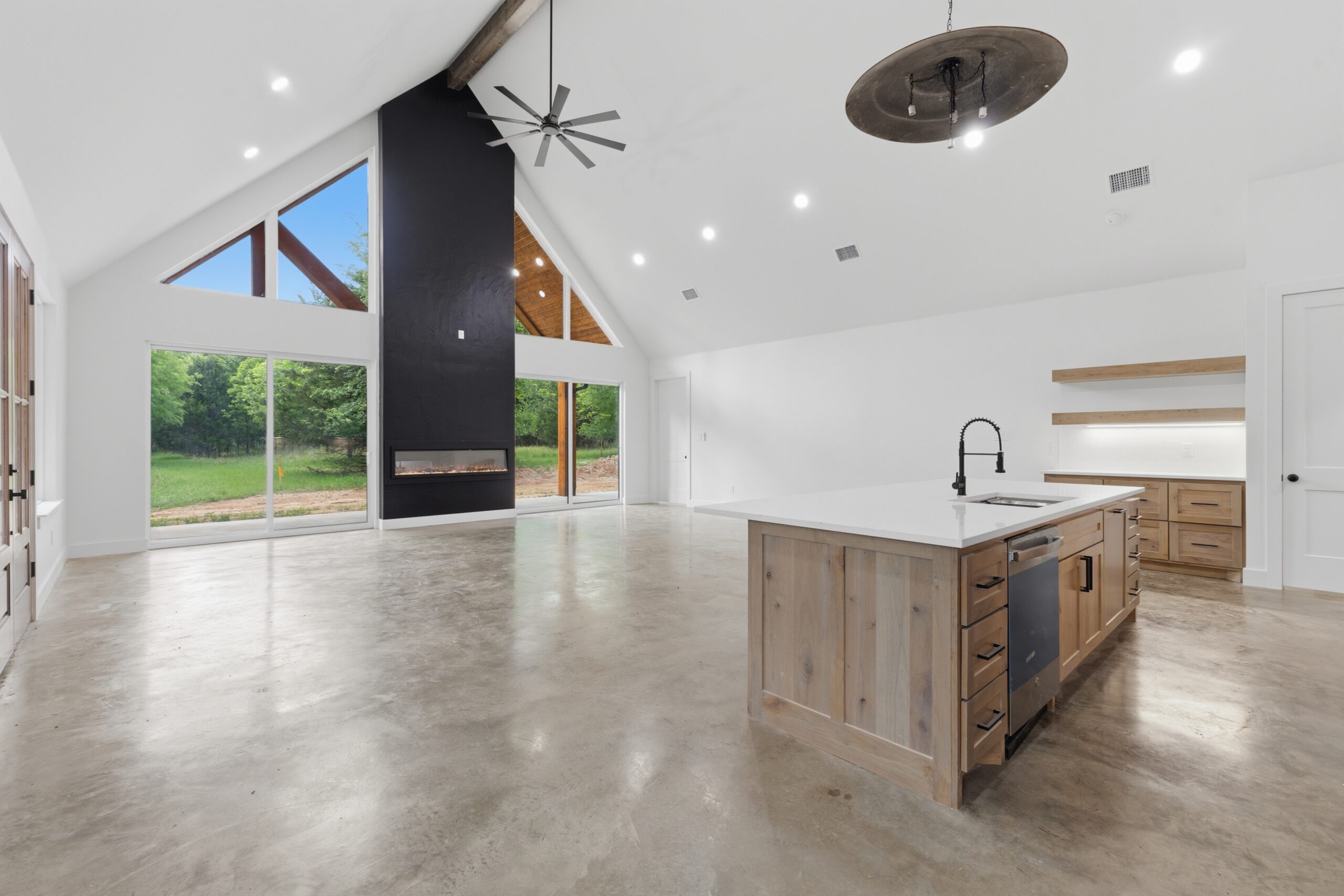 Spacious modern kitchen and living area with vaulted ceiling, large windows, concrete floors, black accent fireplace, and central island with sink.