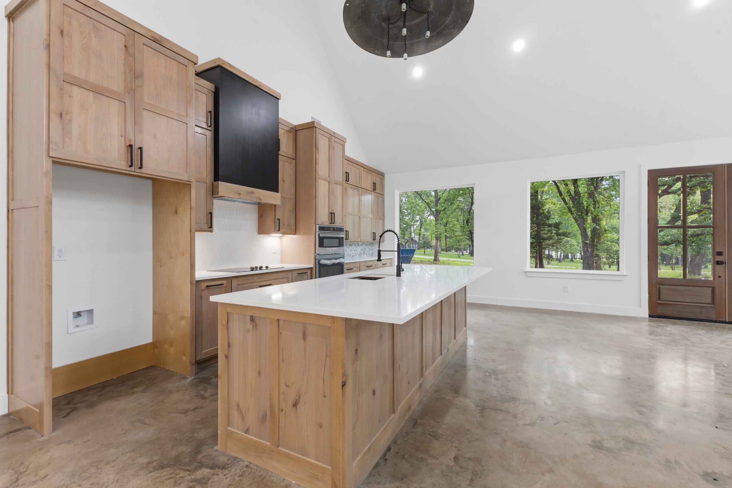 Modern kitchen with wood cabinetry, white countertops, an island with a sink, built-in appliances, large windows, and a glass door overlooking a green outdoor area.