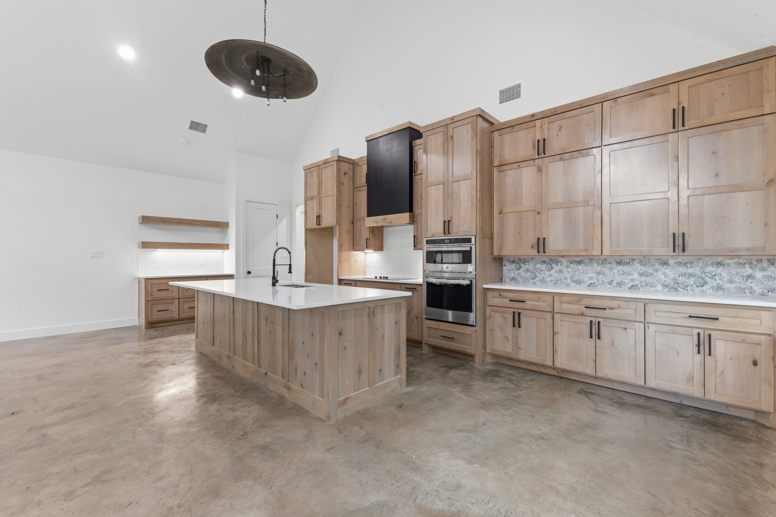 Modern kitchen with wood cabinets, a large island with a sink, stainless steel appliances, and concrete floors. The room features white walls and a patterned tile backsplash.