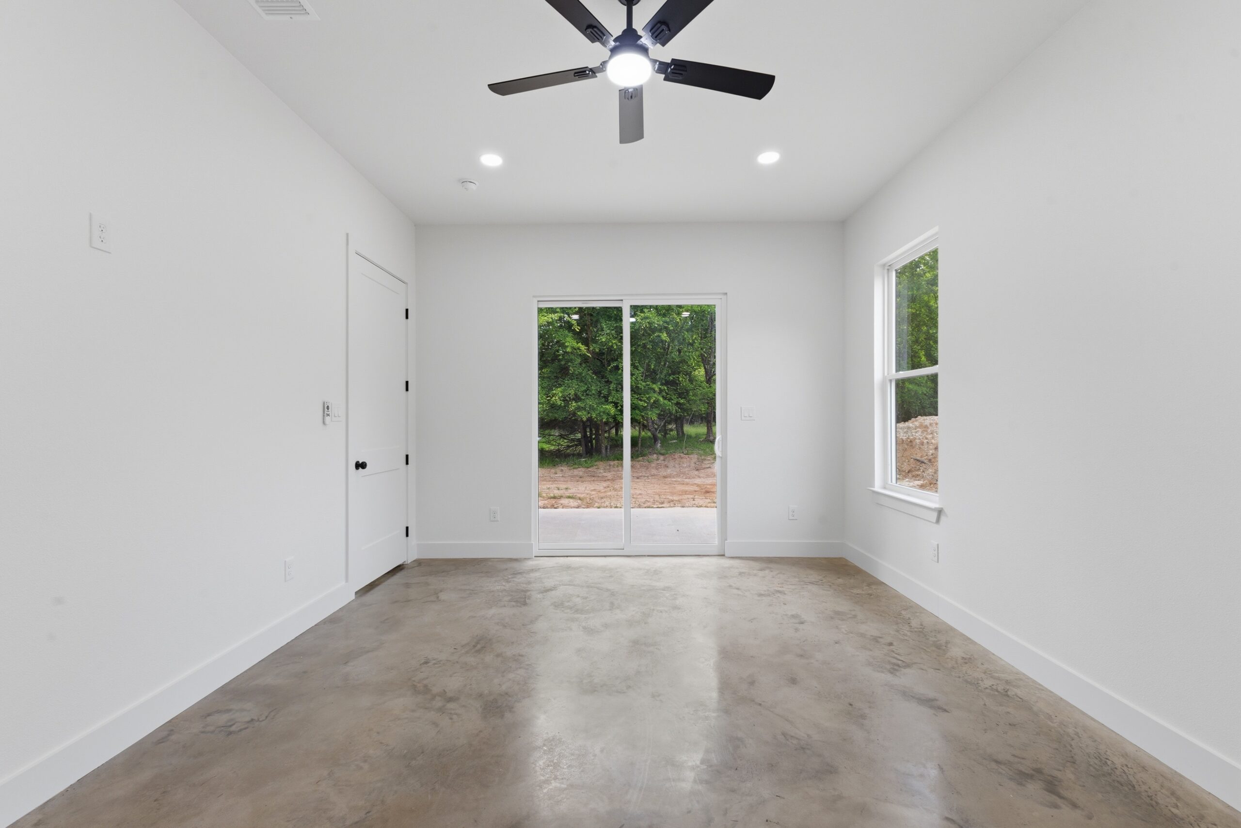 Empty room with white walls, concrete floor, ceiling fan with light, a window, and a glass sliding door leading outside to a yard with trees.