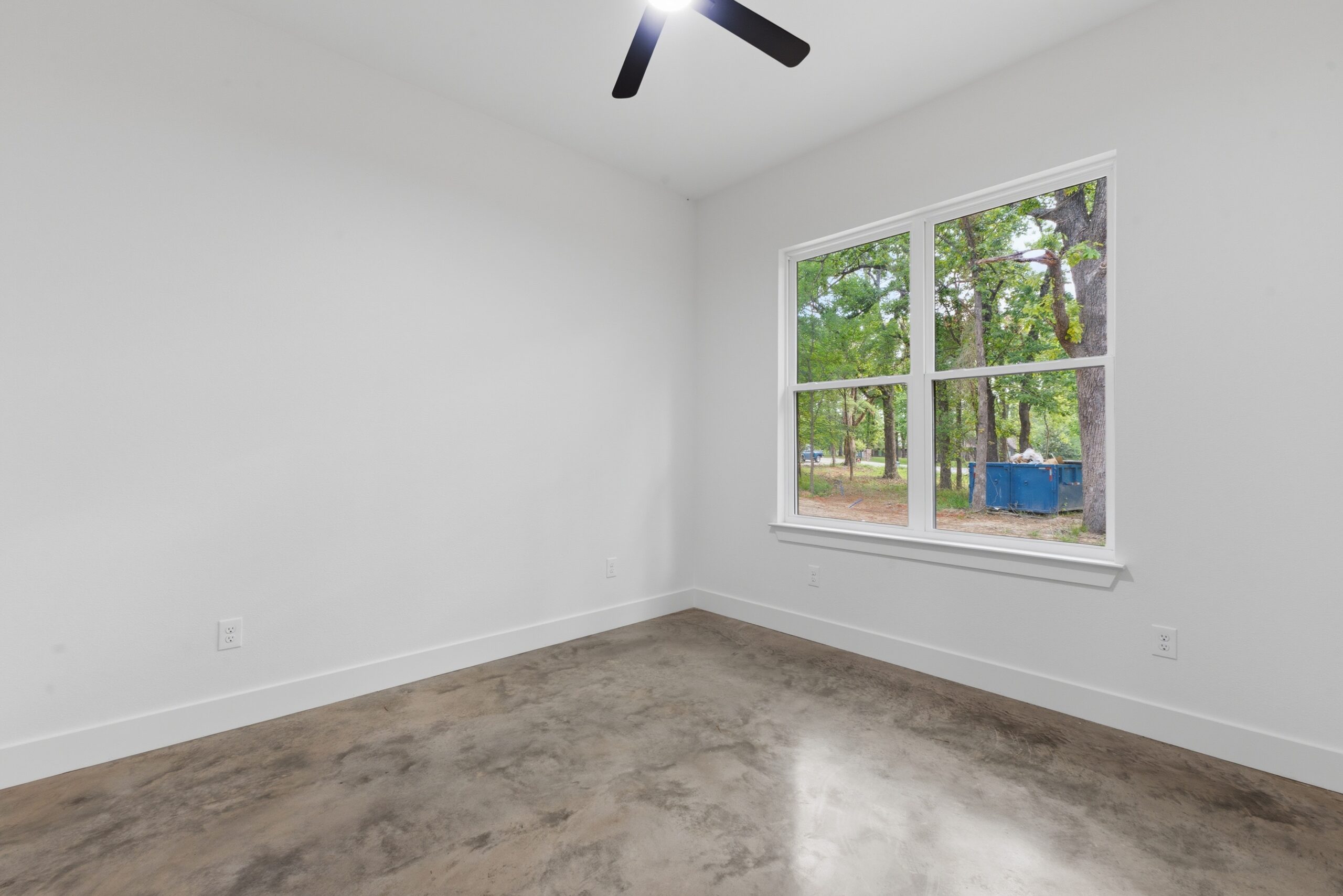 Empty white room with a large window showing a tree-filled outdoor view; polished concrete floor.