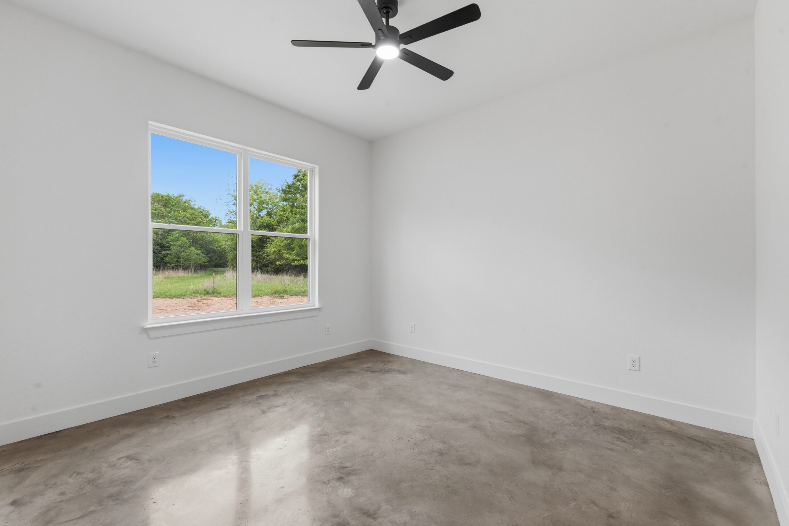 Empty white unfurnished room with a concrete floor, a large window showing green trees outside, and a ceiling fan with a light.
