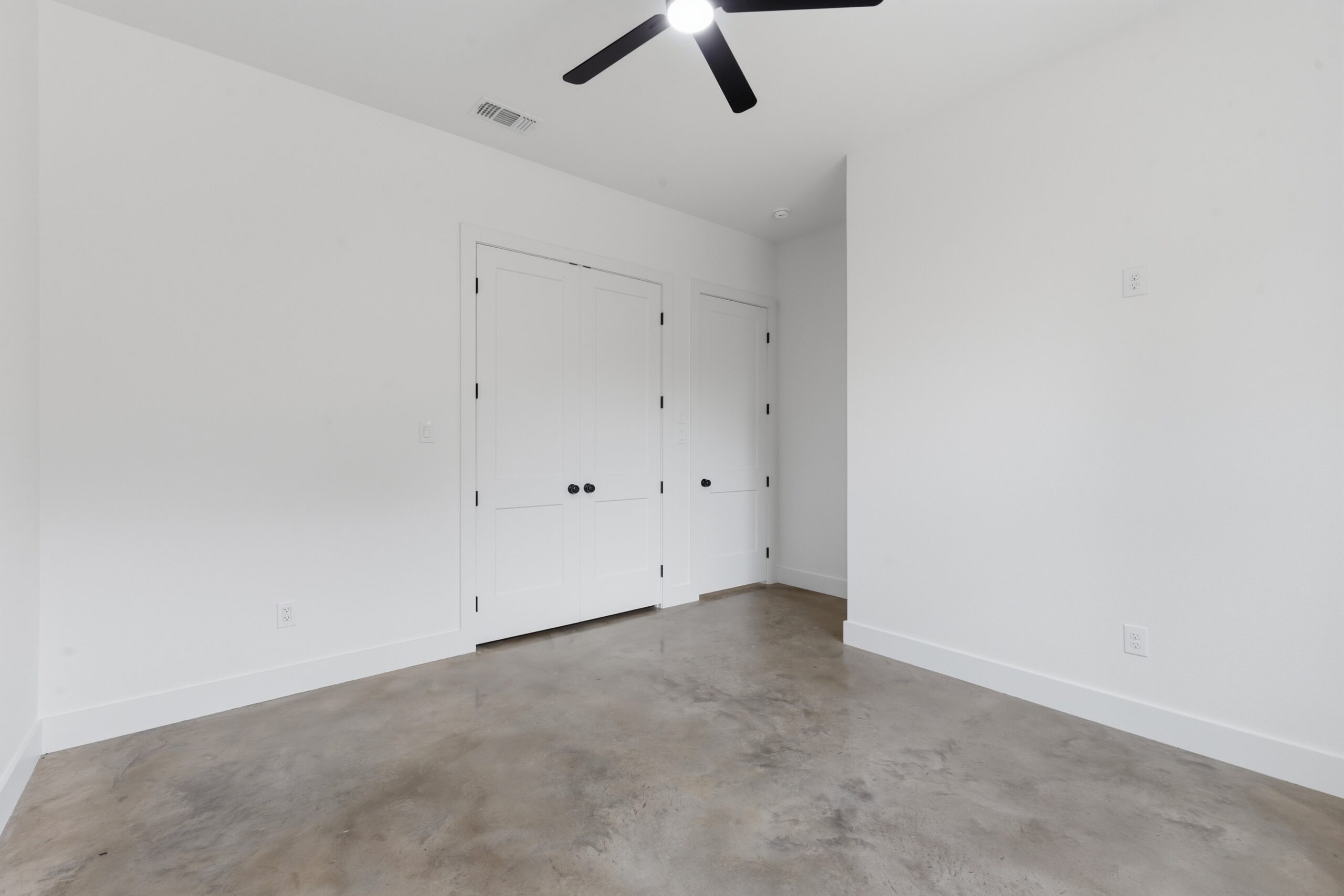 White empty room with a concrete floor, two white paneled doors, and a ceiling fan overhead.