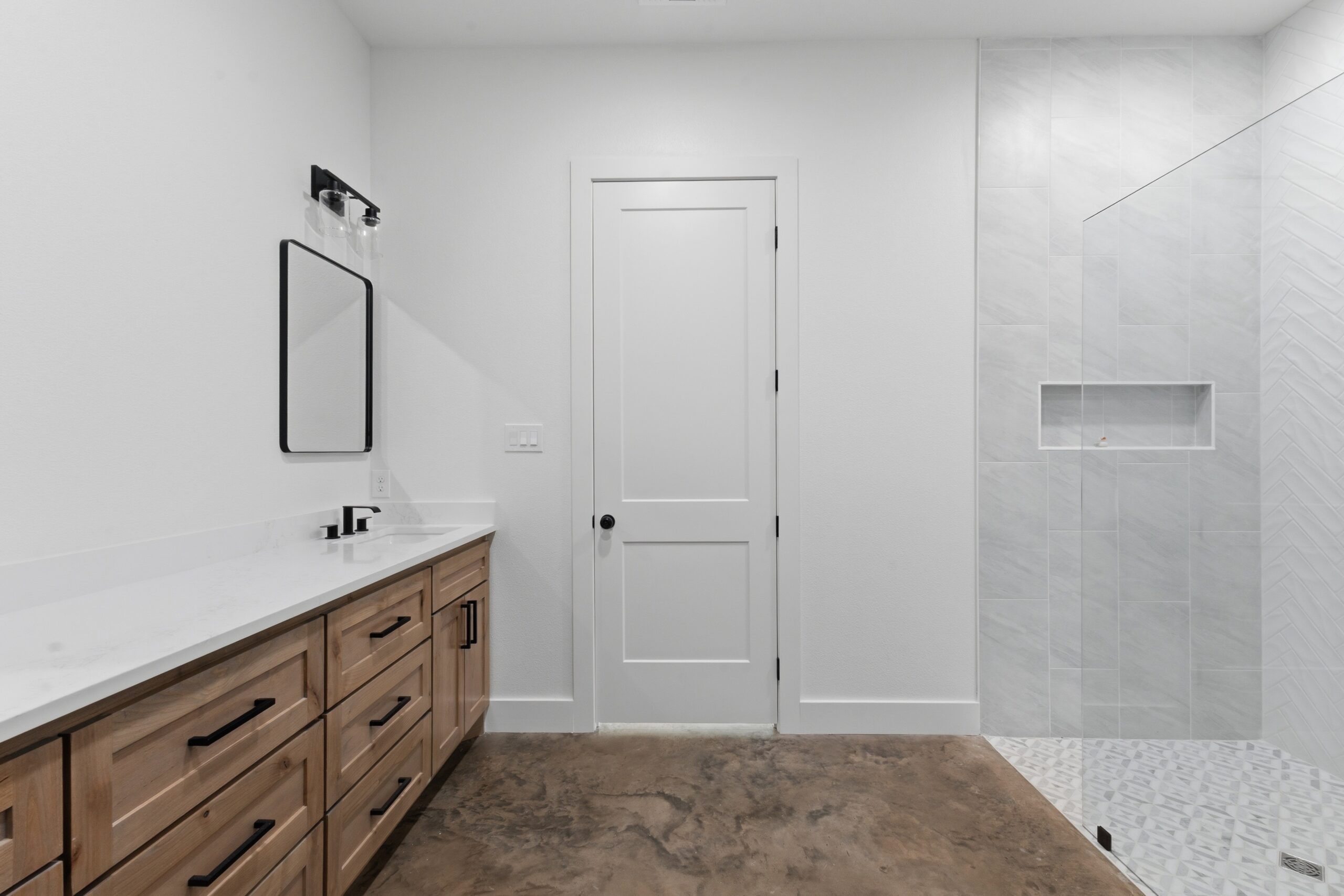 Modern bathroom featuring a wooden vanity with black hardware, a white countertop, and a rectangular wall mirror; center door and a tiled shower area to the right.