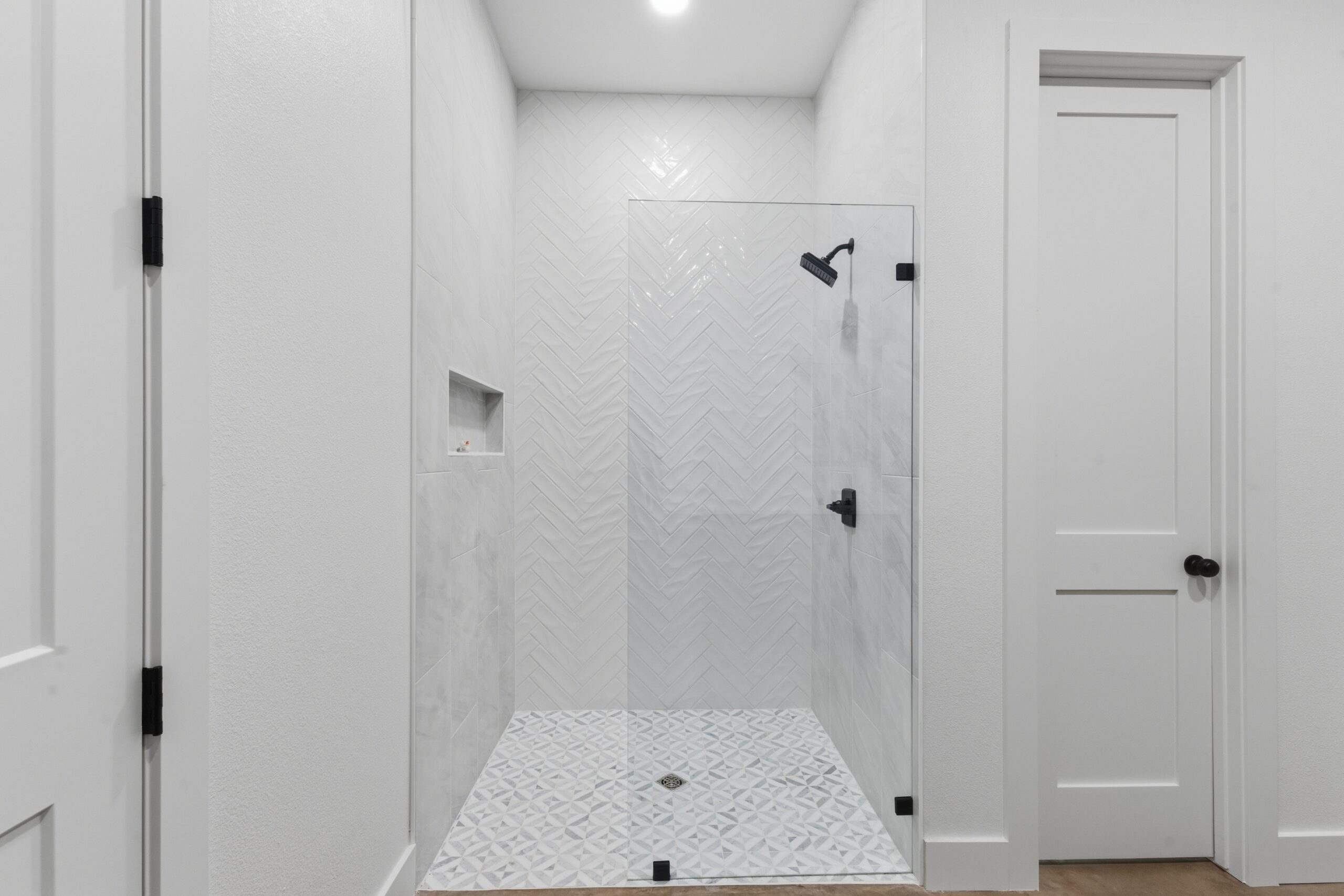 White walk-in shower with herringbone tile walls, glass door, and black fixtures; built-in niche visible on left wall.