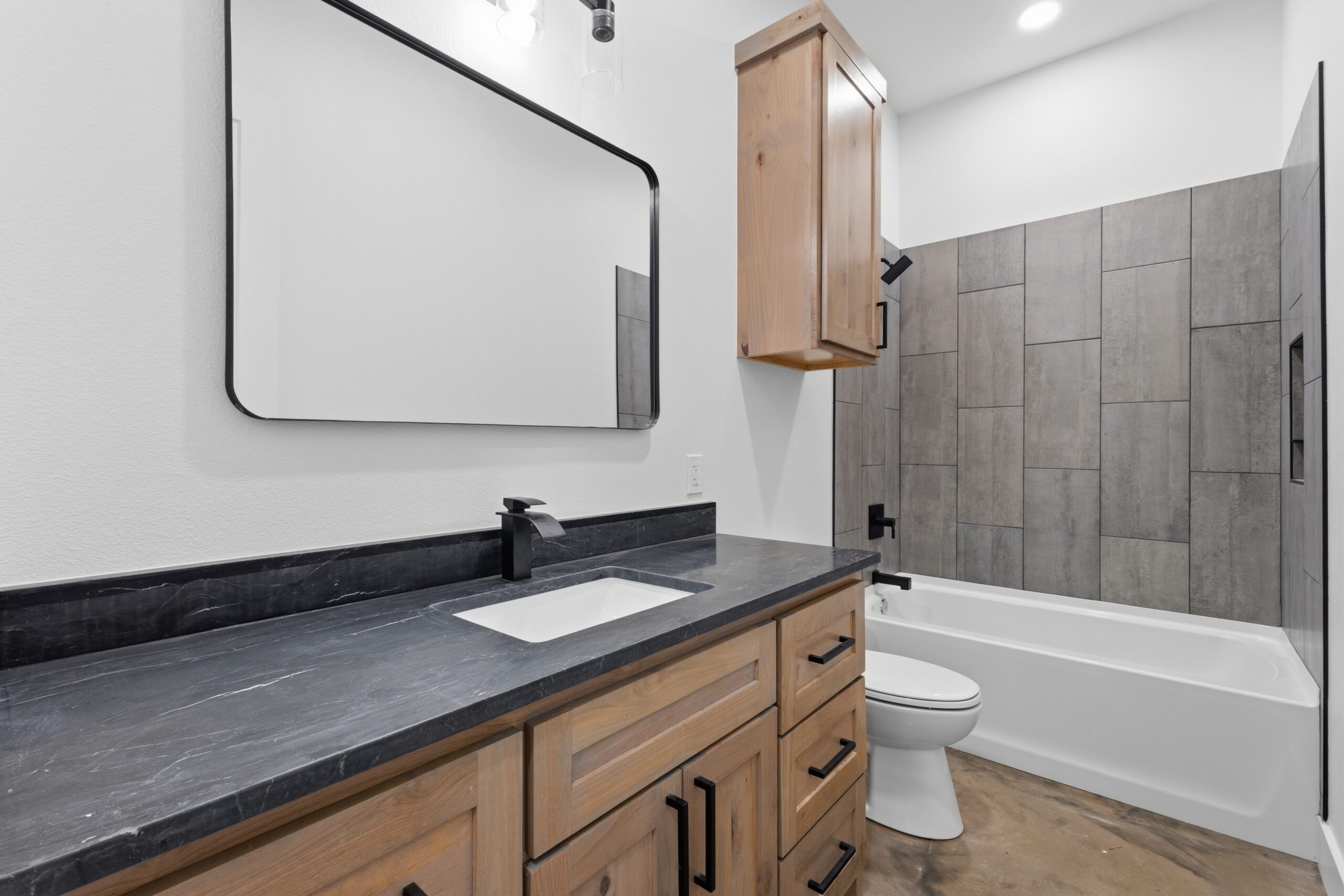 Modern bathroom with a wood vanity, black countertop, rectangular mirror, wall cabinet, toilet, and a bathtub with gray tile surround.