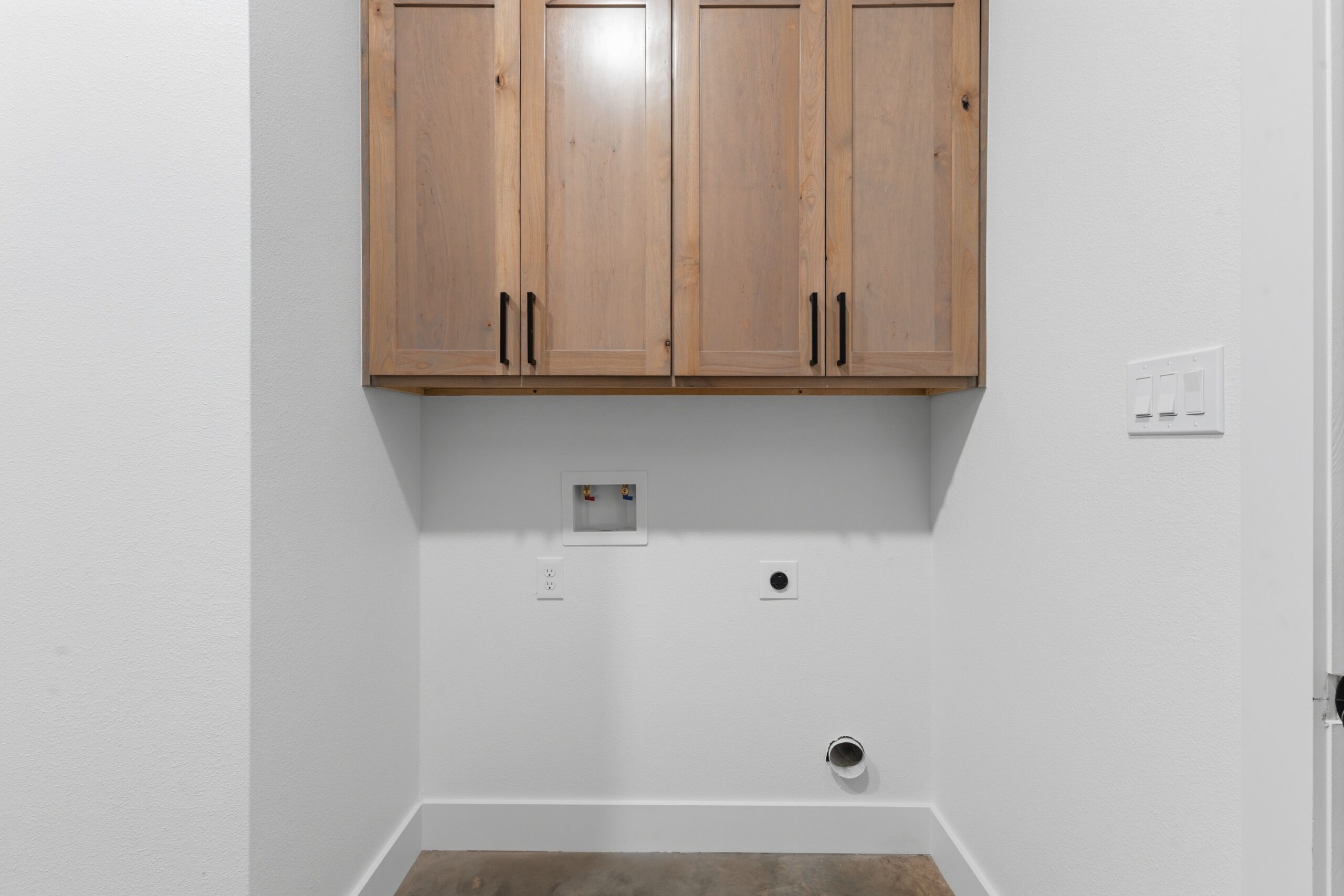 Laundry area with upper wooden cabinets, washer hookups (hot/cold valves), drain box, electrical outlet, and a round dryer vent opening on the wall.
