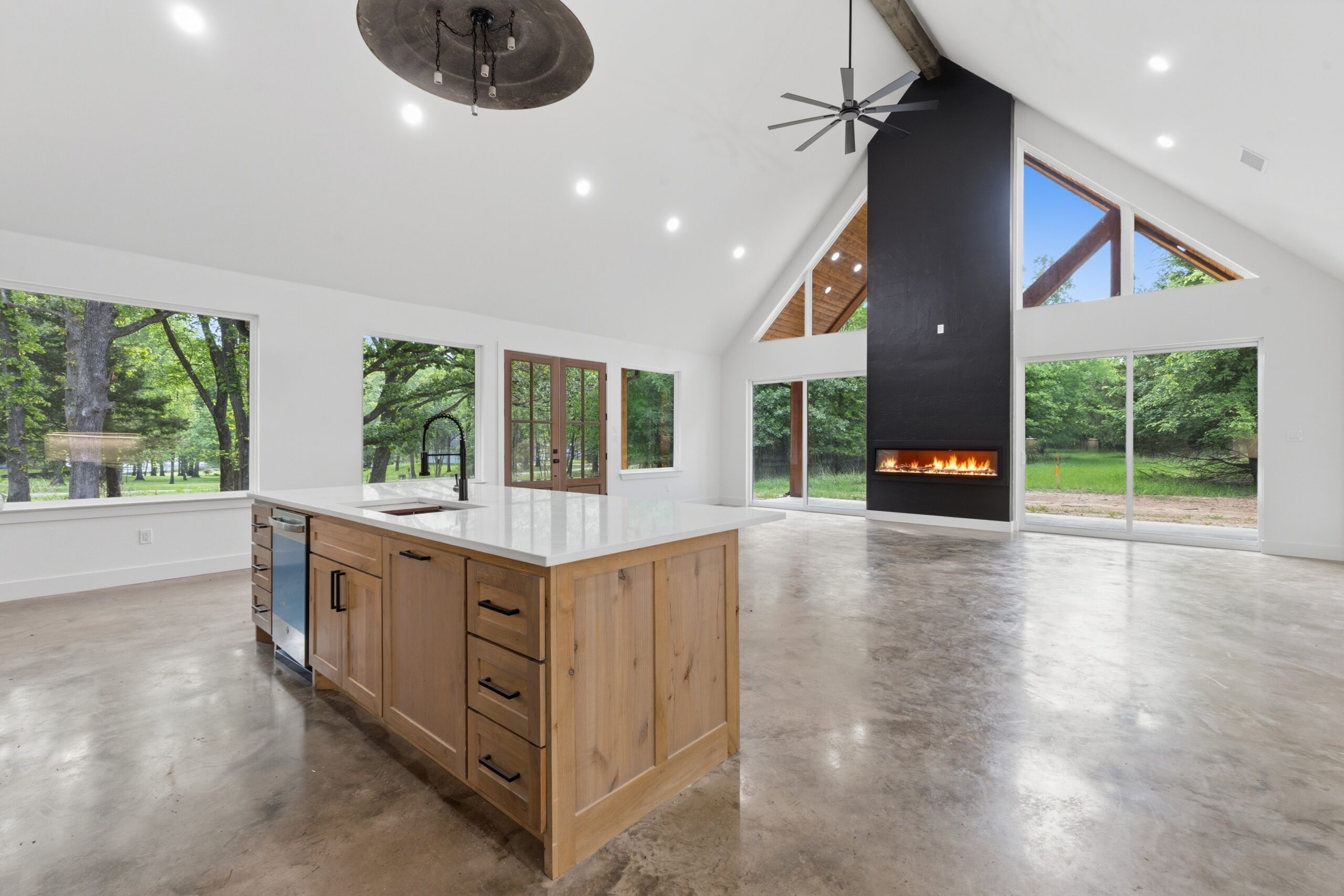 Bright open-concept kitchen and living area with a large wooden island, white counters, and floor-to-ceiling windows showing a green outdoor scene.