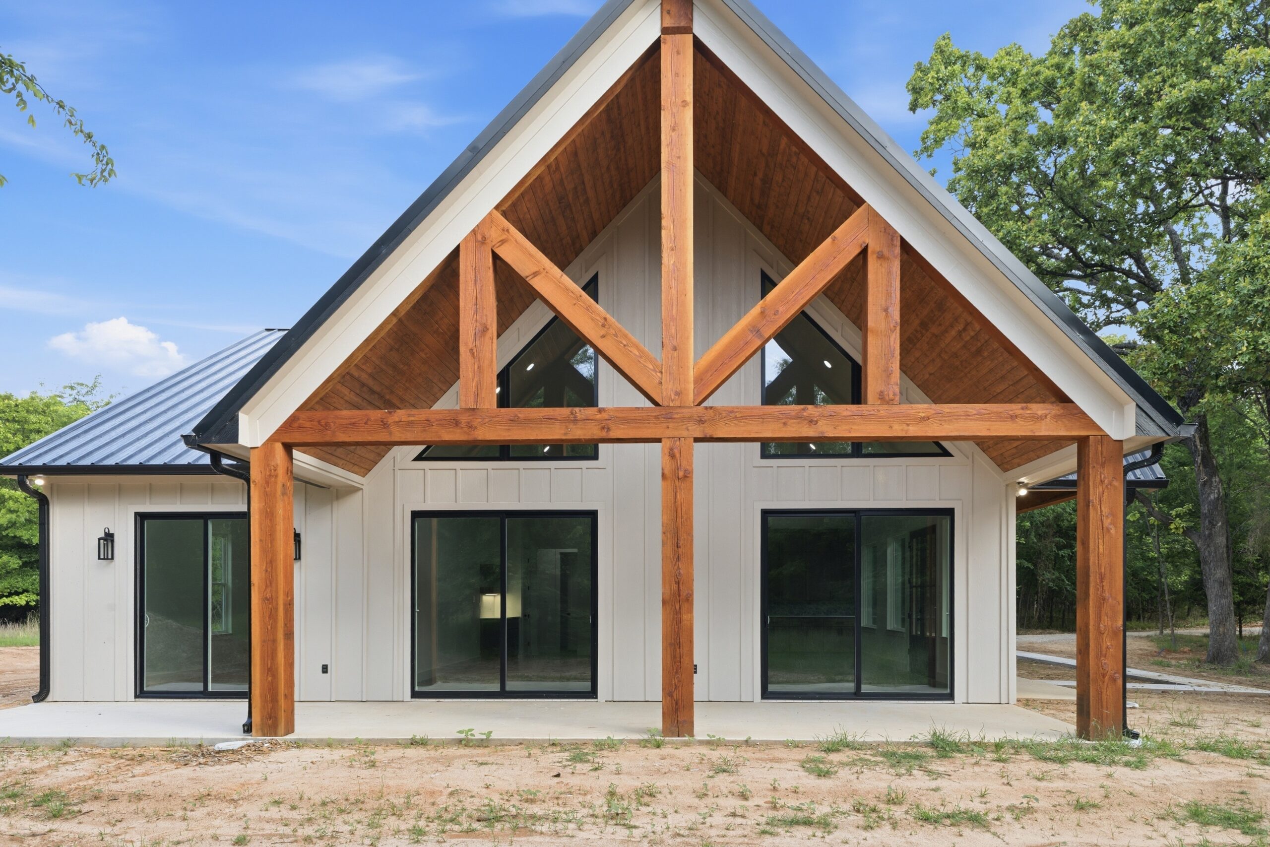 A modern house with a steeply pitched roof, large wooden beams, white siding, and multiple sliding glass doors, surrounded by trees.