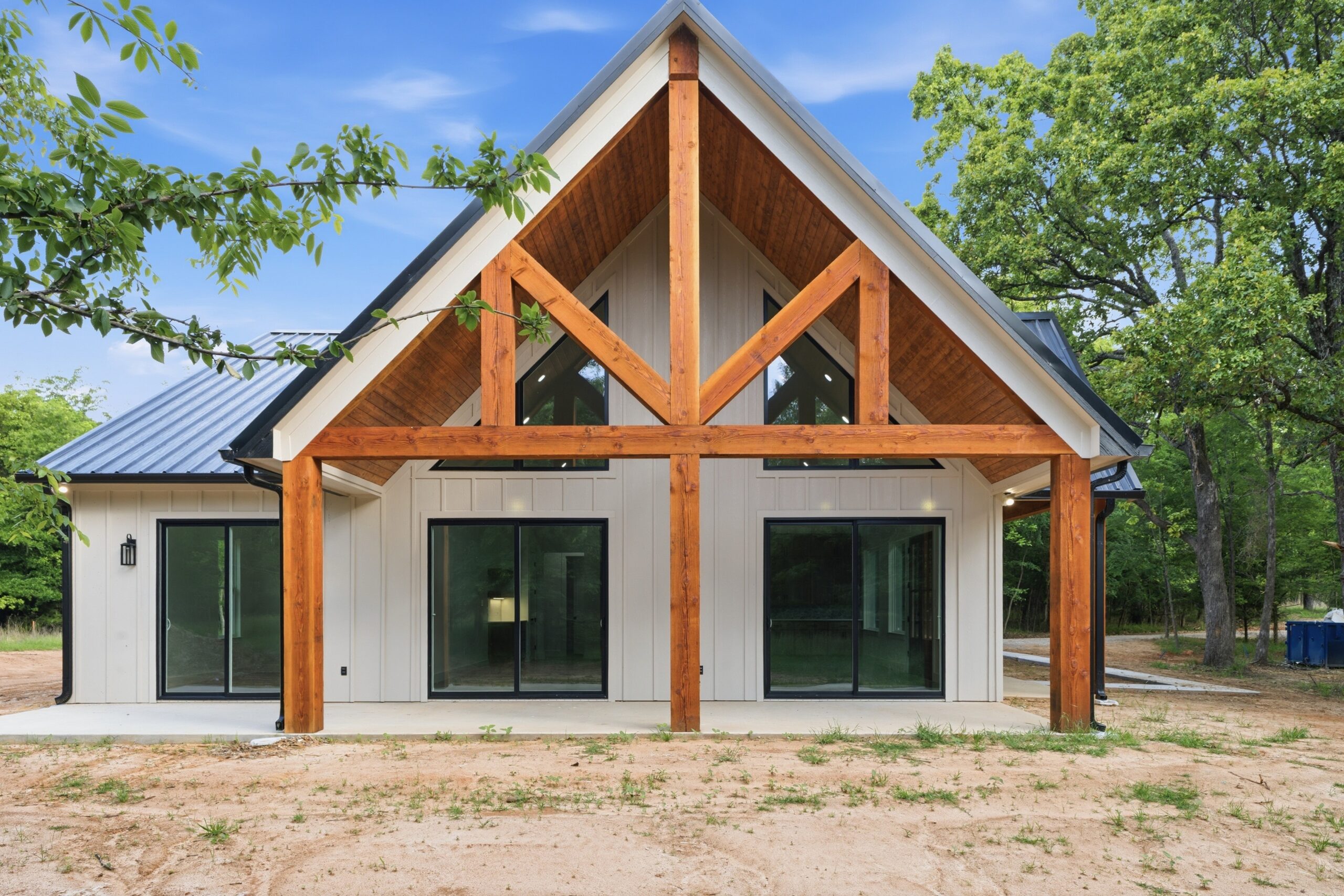 Modern house exterior with a steep gable roof, exposed wooden beams, large windows, and white siding, surrounded by trees and a dirt yard.