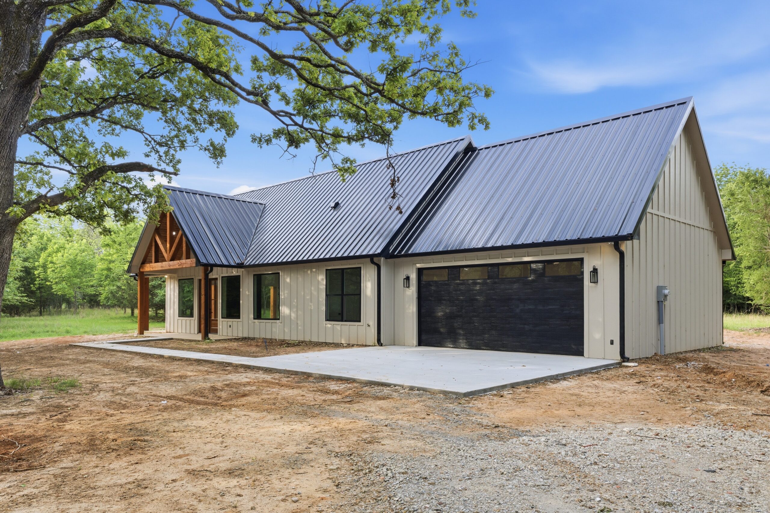 Modern beige house with a steep dark metal roof under a large leafy tree, in a rural yard