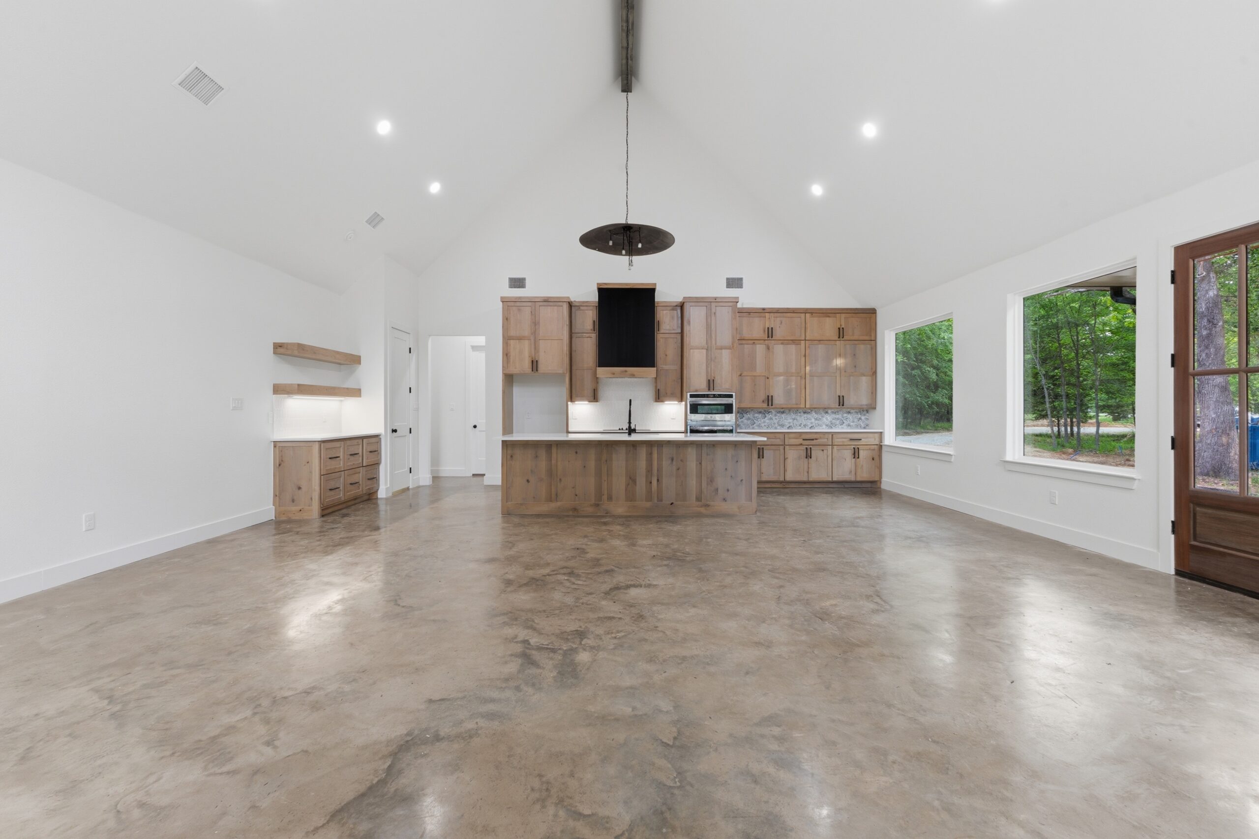 Spacious modern kitchen with wooden cabinets, center island, stainless steel appliances, and large windows overlooking greenery, featuring a polished concrete floor.