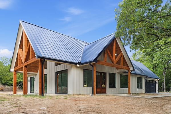 Modern house with a metal gable roof, large wooden support beams, and white vertical siding, situated on bare ground with trees and blue sky in the background.