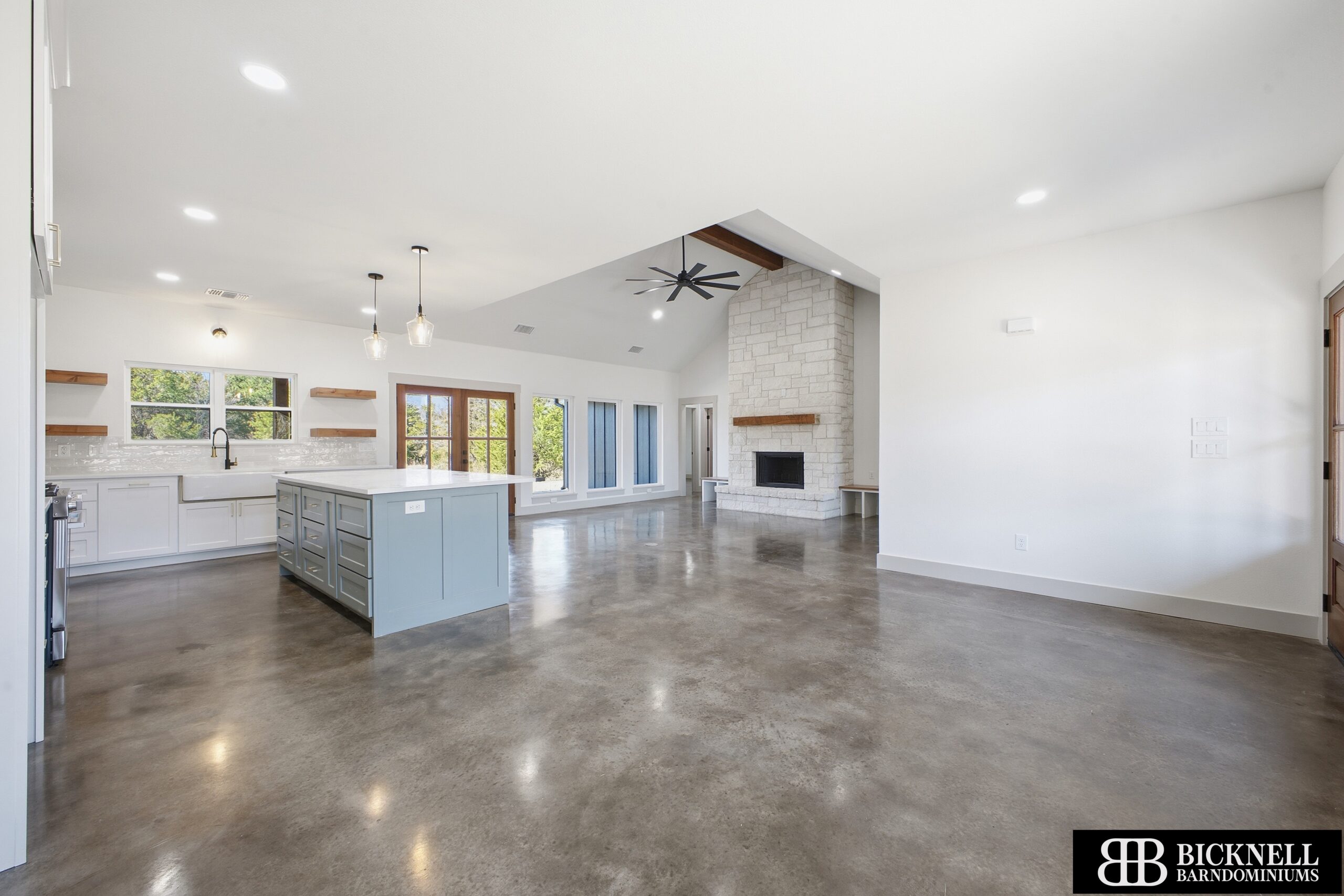 Open-concept kitchen with a blue island, white cabinets, pendant lights, and large windows showing greenery outside.