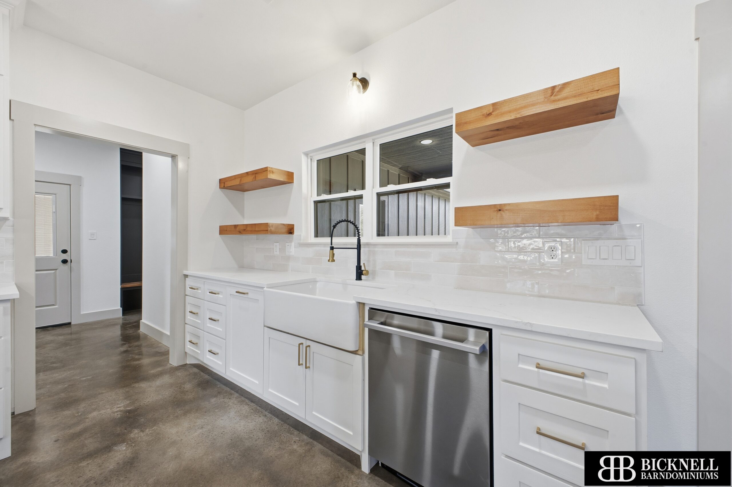 Modern kitchen with white cabinets, farmhouse sink, stainless steel dishwasher, wooden floating shelves, and polished concrete floor. A window is above the sink.
