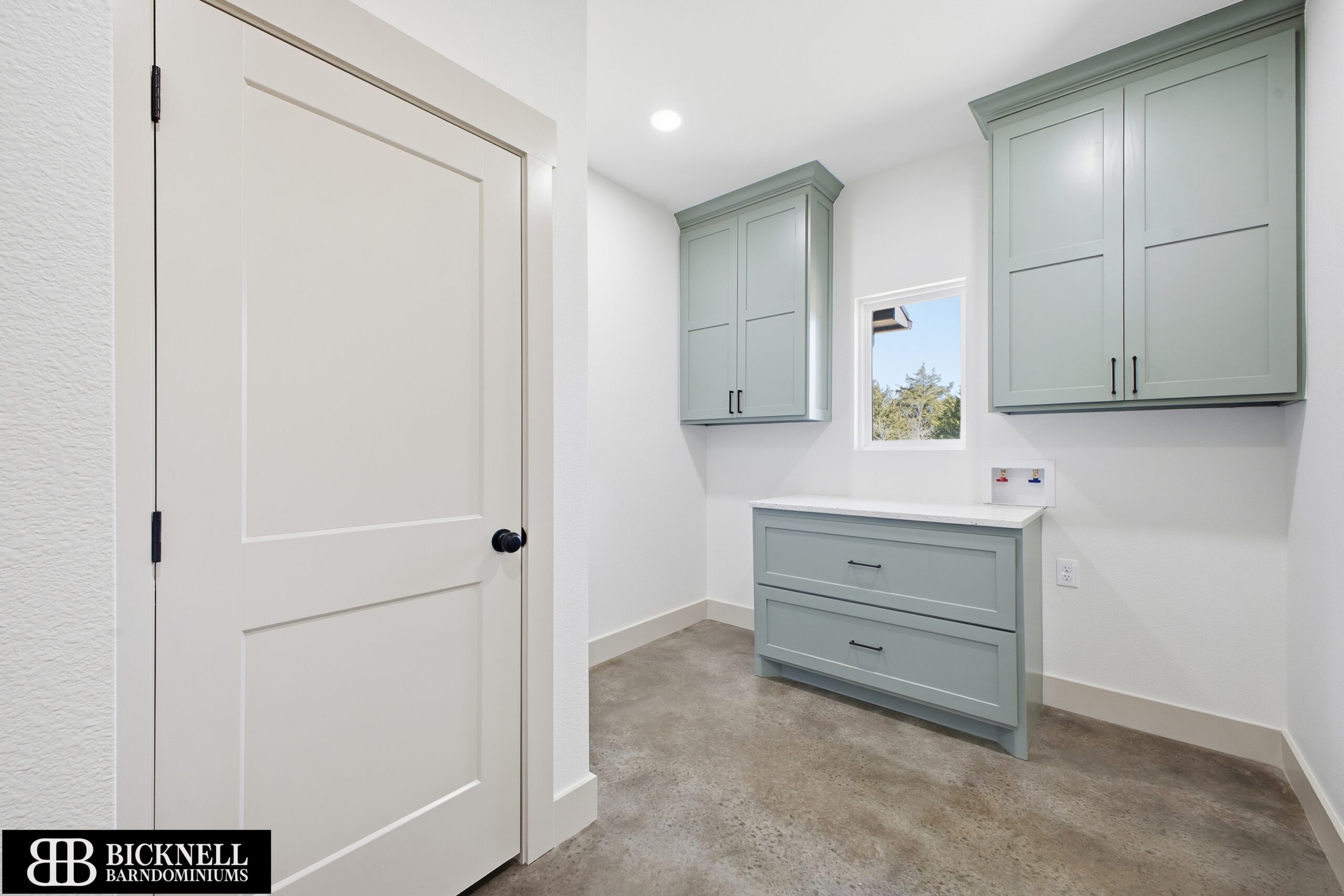 Laundry room with sage green upper and base cabinets, white walls, concrete-look floor, and a small window above a countertop with hookups.