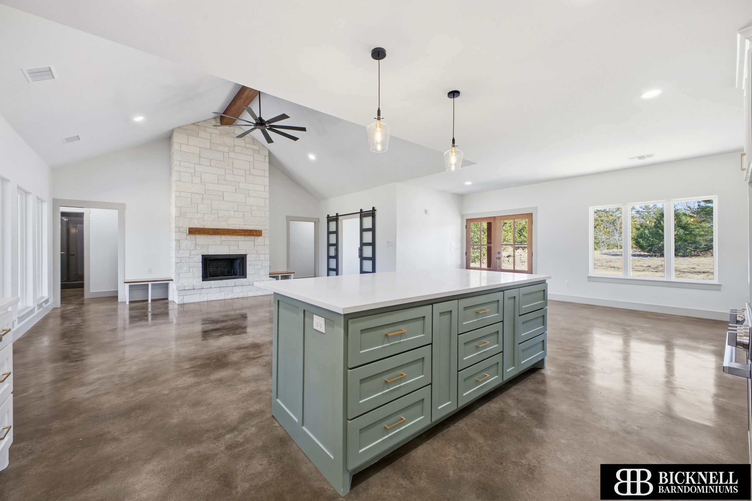 Open-concept kitchen with a large green island, white countertops, and a stone fireplace, under a vaulted ceiling with pendant lights and a ceiling fan.