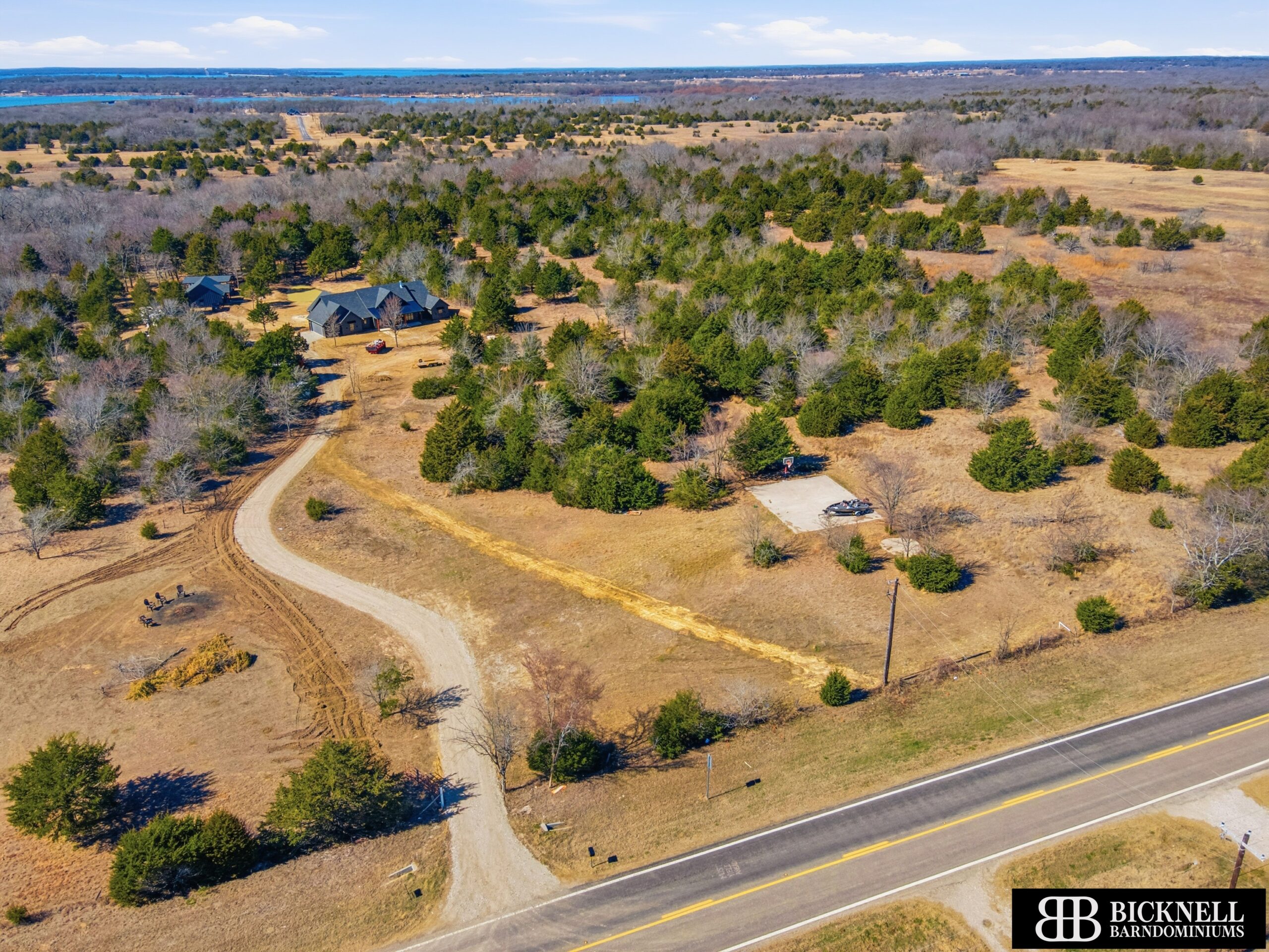 Aerial view of a rural property with a winding driveway, scattered trees, a house, outbuildings, and nearby road, with open fields and water visible in the distance.