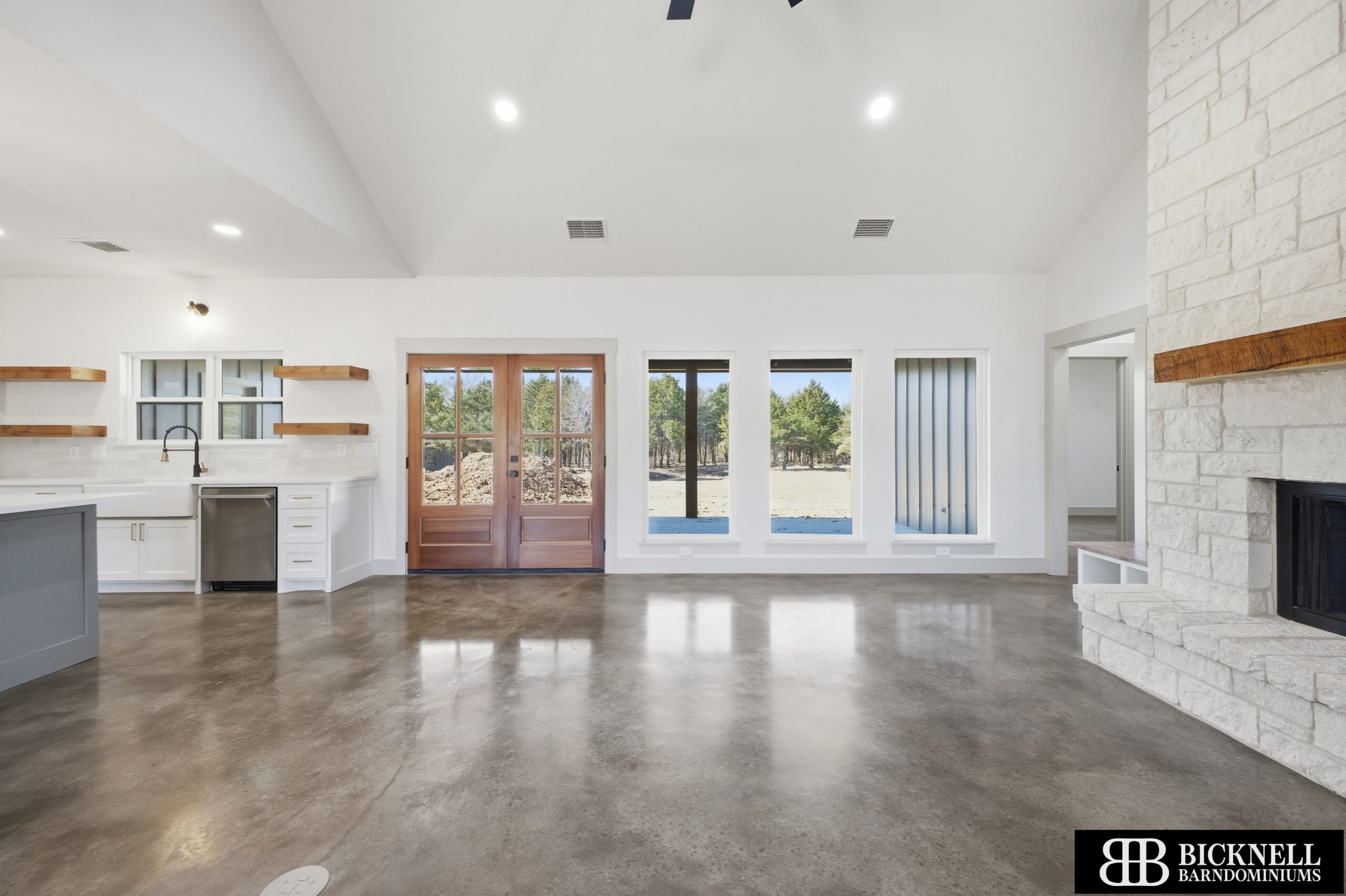 Bright open-plan kitchen with white cabinets, wood shelves, and double wooden doors opening to outdoors; stone fireplace to the right.