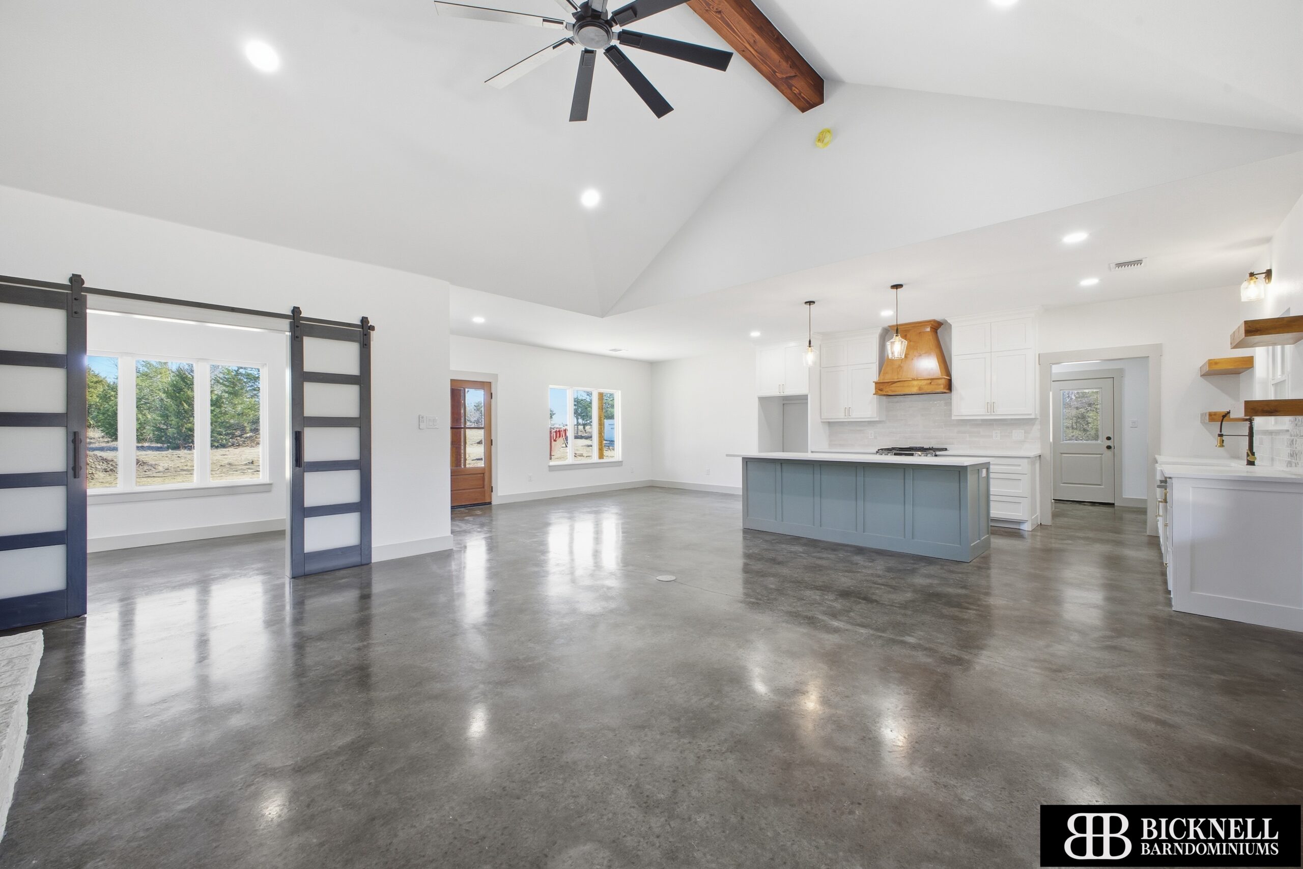 Modern open-concept living area with polished concrete floors, sliding barn doors, and a kitchen featuring white cabinets and a blue island under pendant lights.