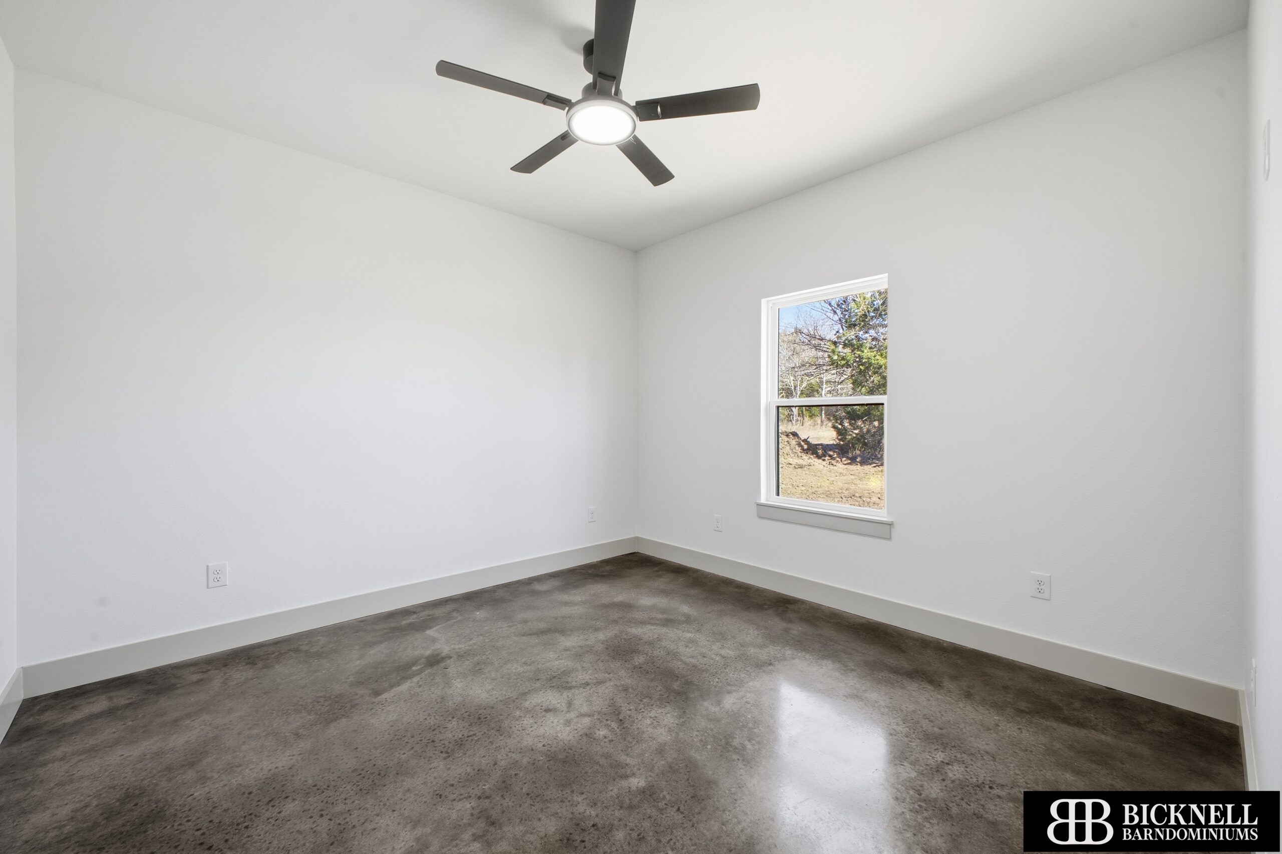 Empty room with white walls, a single window, polished concrete floor, and a ceiling fan with a light. The logo "Bicknell Barondominiums" is in the bottom right corner.
