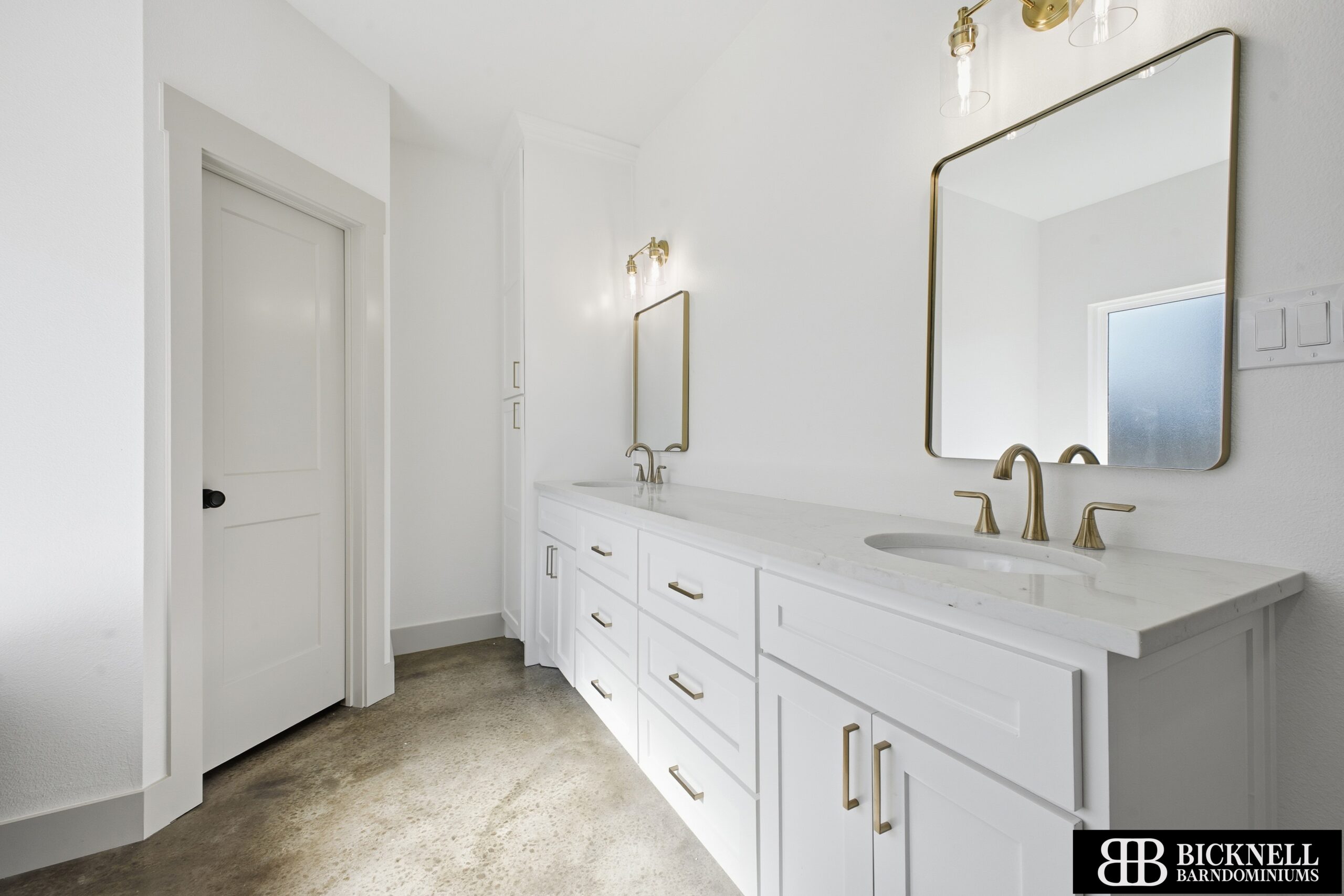 A modern bathroom with white cabinets, double sinks, gold fixtures, large mirrors, and concrete flooring.