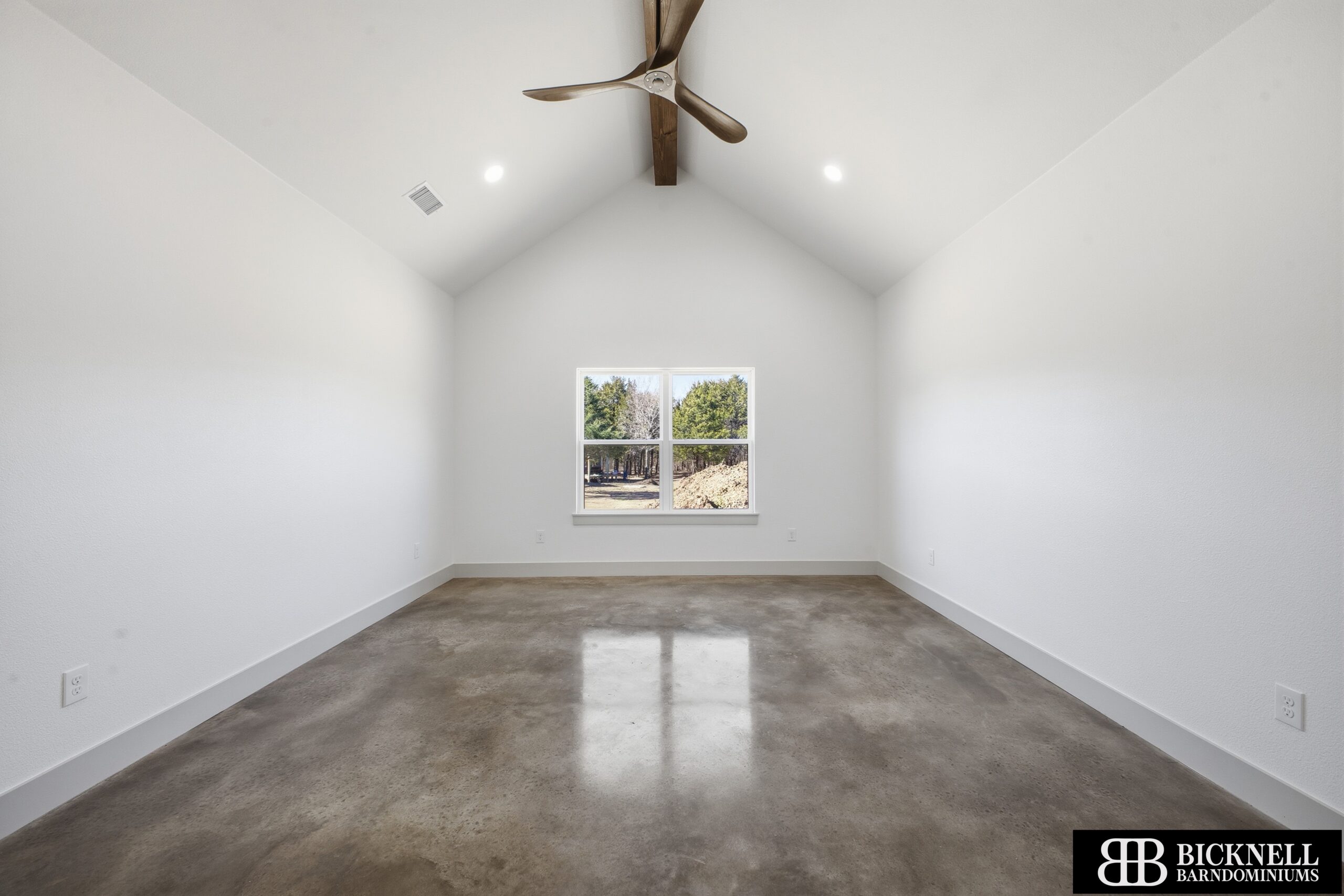 Empty bright room with white walls and a vaulted ceiling; a wooden ceiling fan hangs from the center and a window looks out to trees outside.