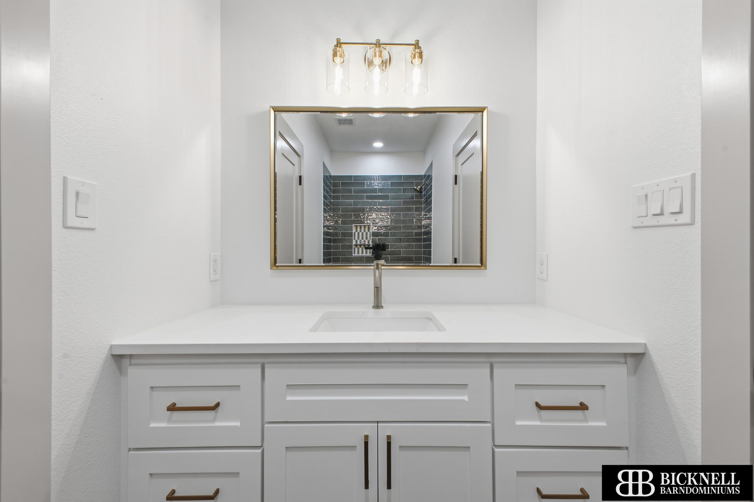 A modern white bathroom vanity with a rectangular mirror, single faucet, and three-bulb light fixture above. Black tile shower reflected in the mirror. Bicknell Barndominiums logo in corner.