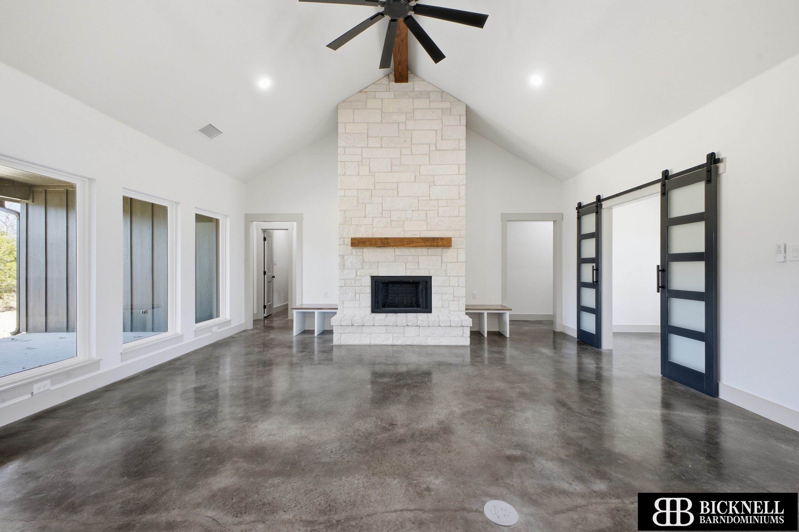 Spacious living room with polished concrete floors, a central stone fireplace, ceiling fan, large windows, and sliding glass doors. Bicknell Barndominiums logo in the corner.