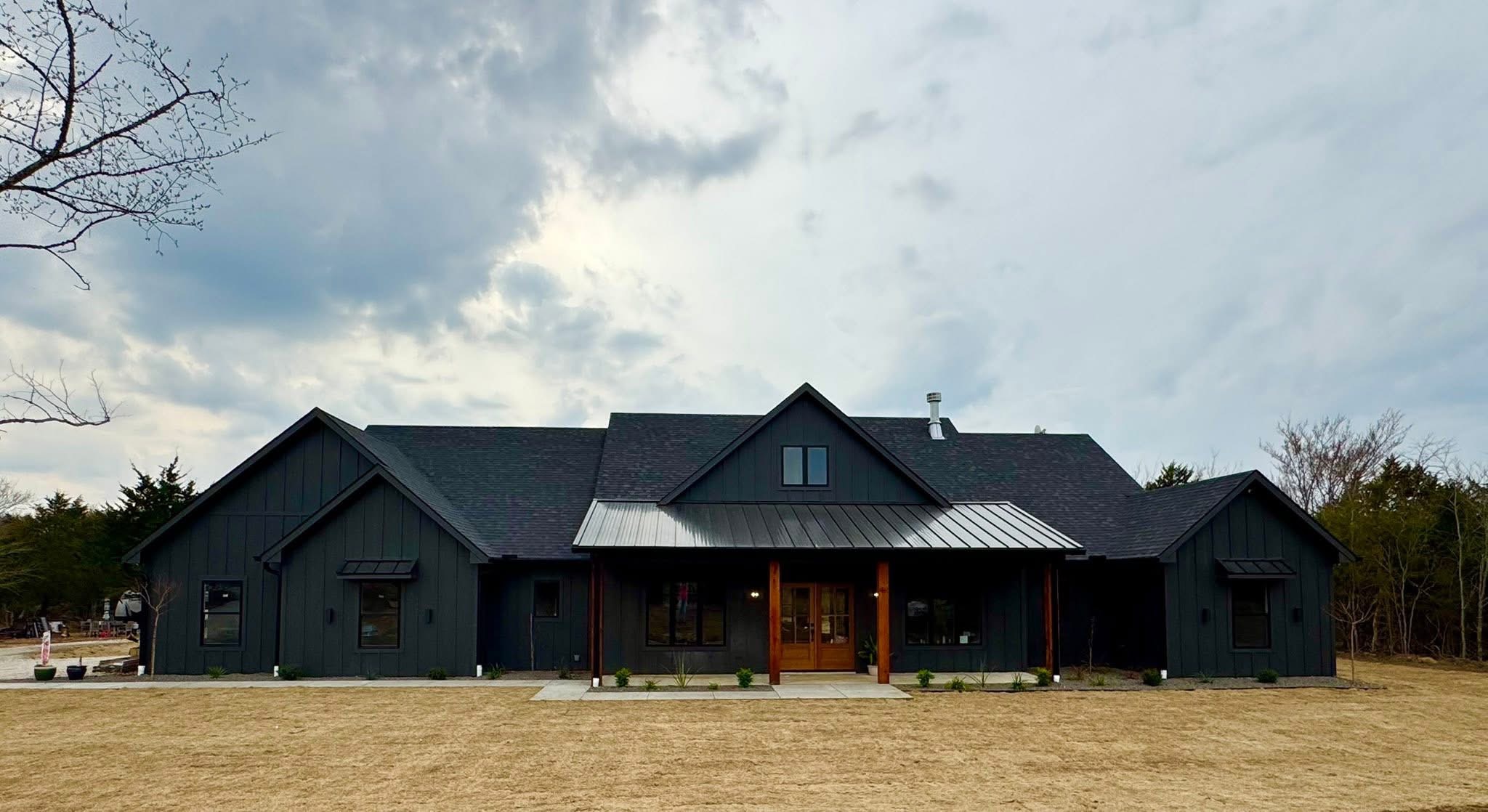 Front view of a dark gray modern house with a peaked roof, central dormer window, and a wooden front porch under a cloudy sky.",