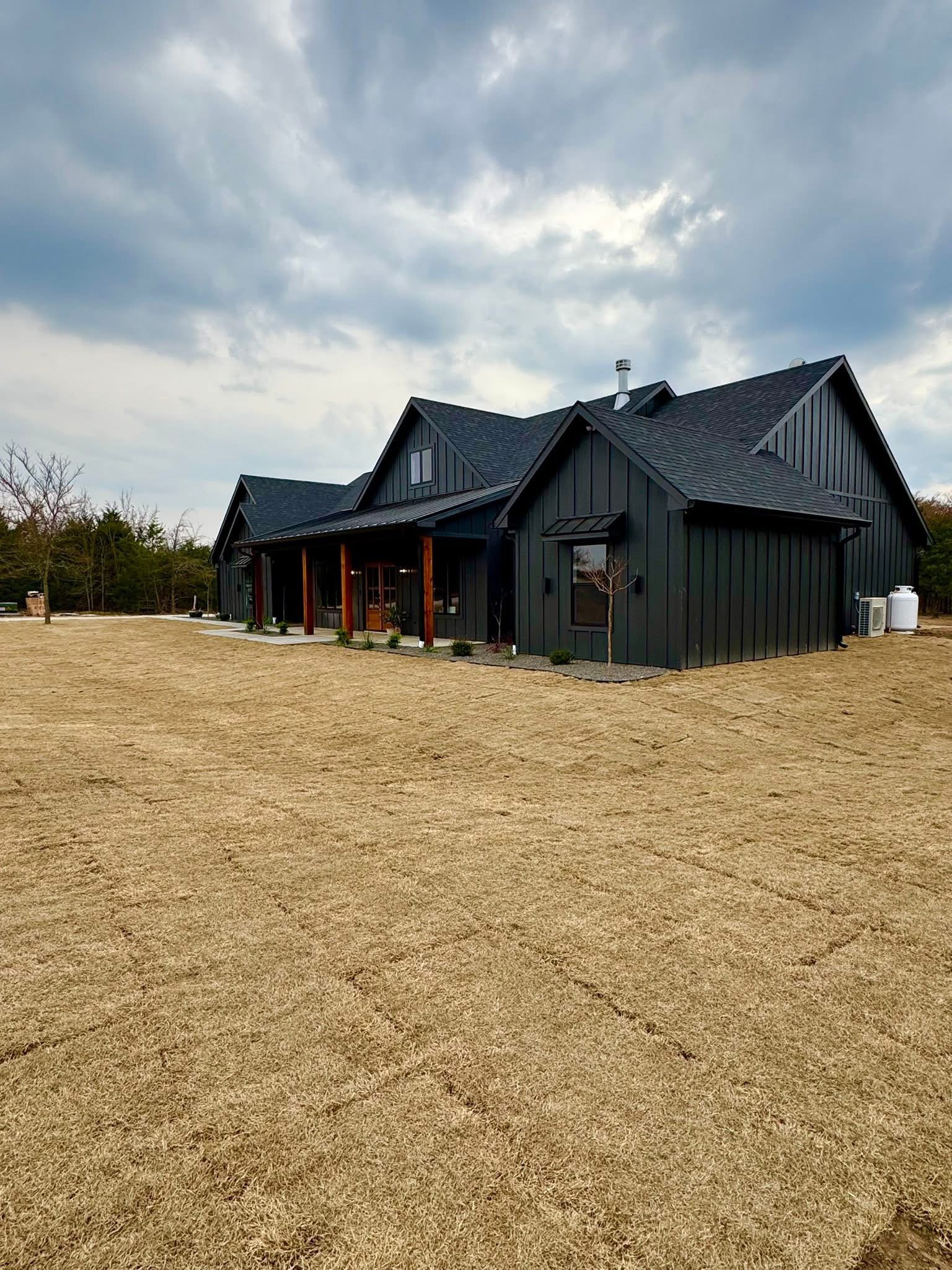 A modern black house with a pitched roof and wooden columns stands on a large, freshly sodded lawn under a cloudy sky.