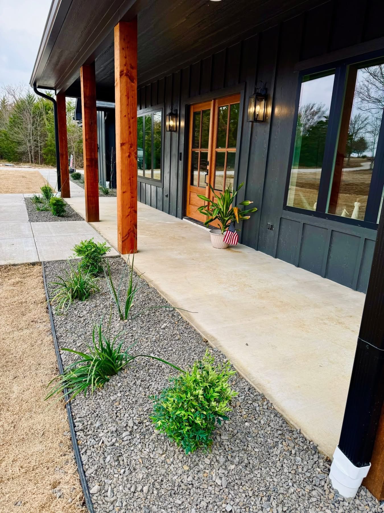 Modern house exterior with wooden pillars, black siding, glass double doors, a potted plant, and landscaped garden bed with small shrubs and gravel along a concrete walkway.
