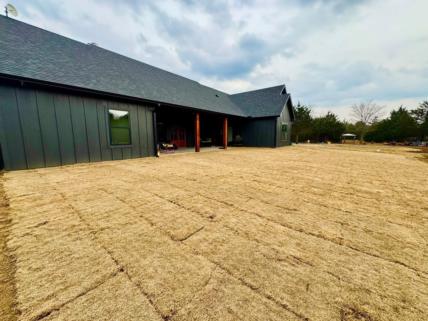 A backyard with freshly laid sod in front of a modern dark-painted house under a cloudy sky.