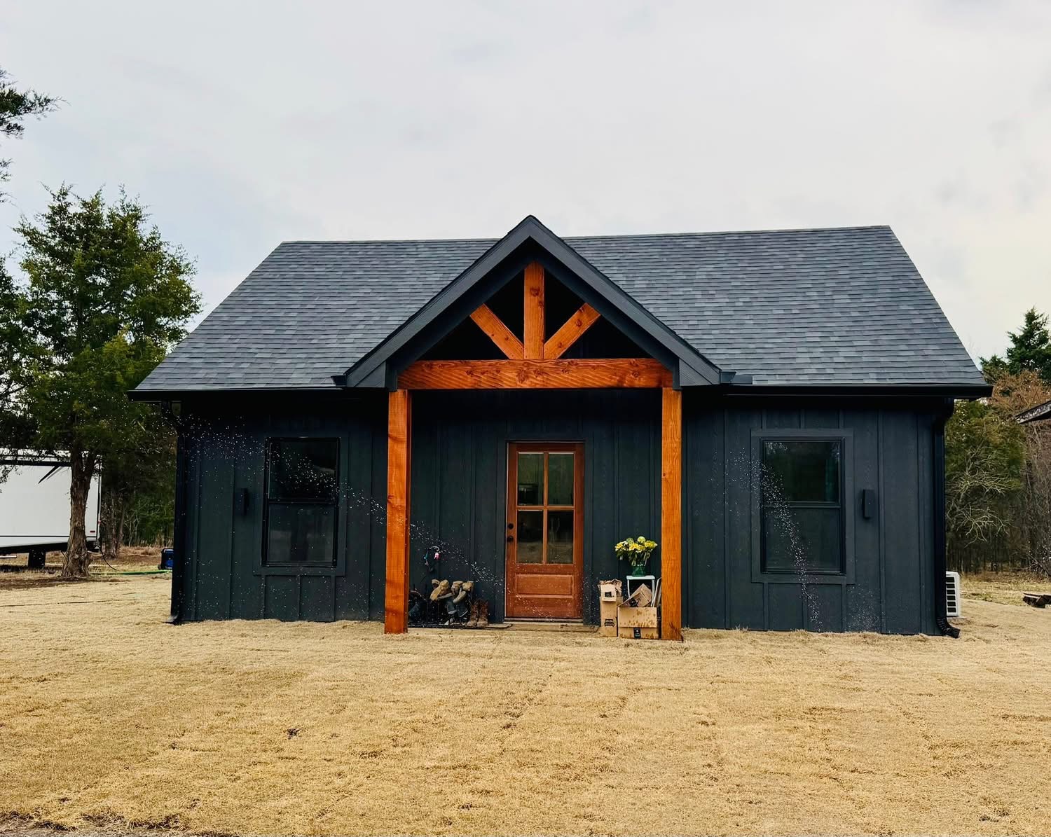 A small, modern dark gray house with a wooden front porch, a central door, and a peaked roof, surrounded by trees and a dry, grassy yard.