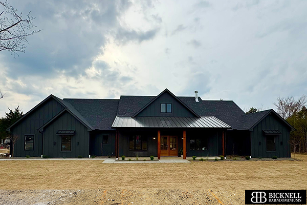 A modern dark-colored barndominium with a gabled roof, large front porch, and wooden pillars, situated on a dry grassy lot under a cloudy sky.